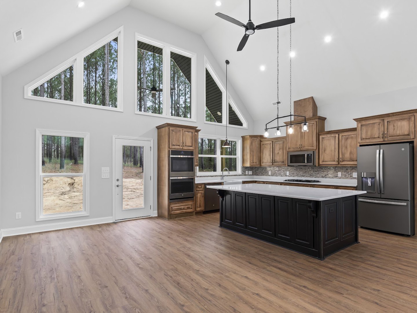 Spacious kitchen featuring a large white island, wood flooring, stainless steel refrigerator, ceiling fan with light, open microwave, and door revealing forest view