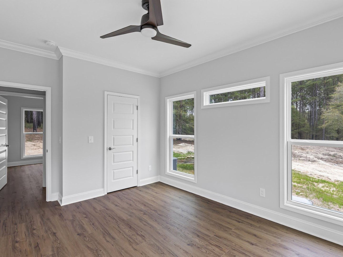 White walls, wood flooring, ceiling fan with light, white door with silver handle, window overlooking trees and a dirt road