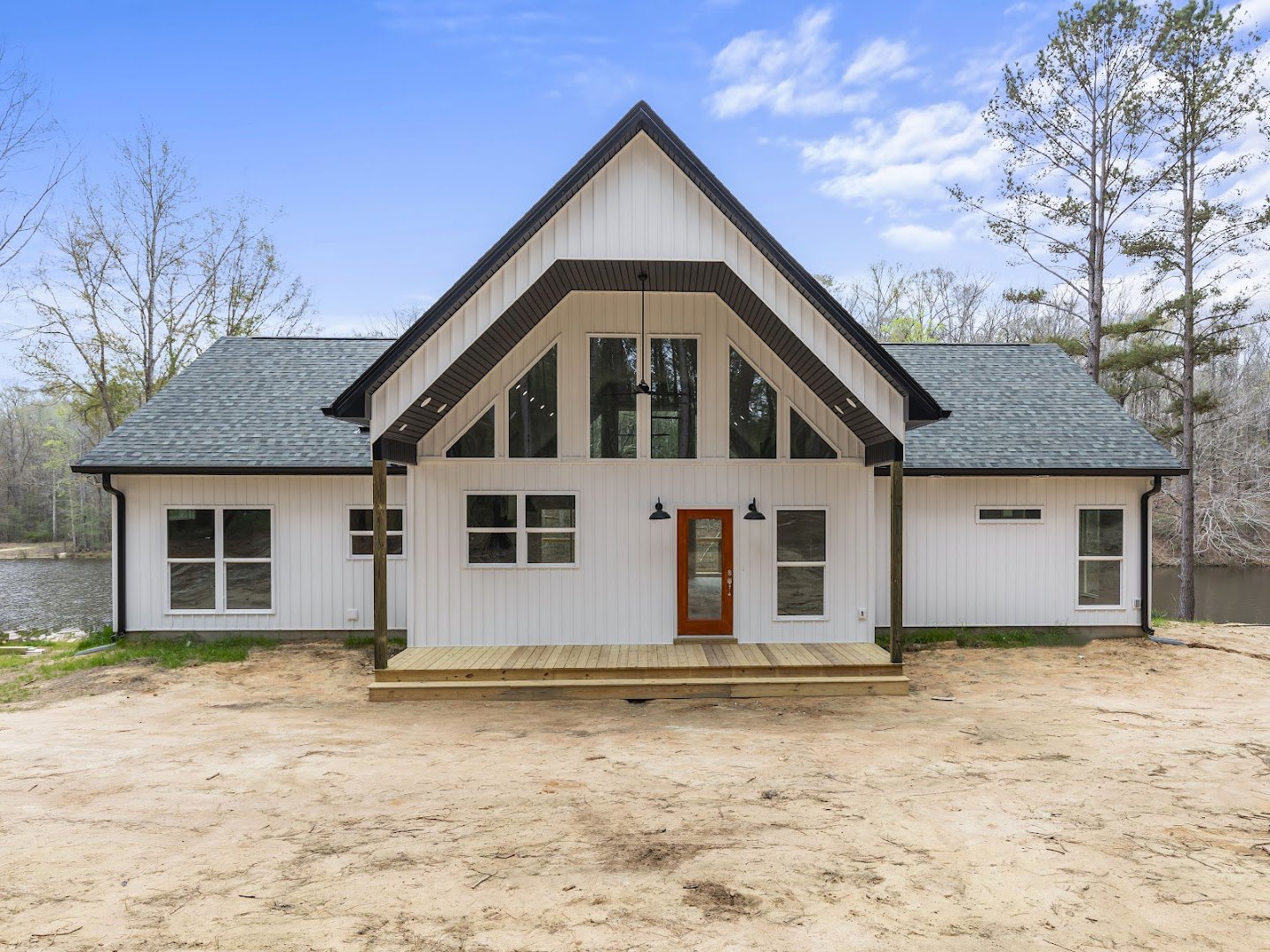 White siding house with wooden porch, glass door, and white-framed windows; wooden deck extends over dirt ground, surrounded by trees and cloudy sky.