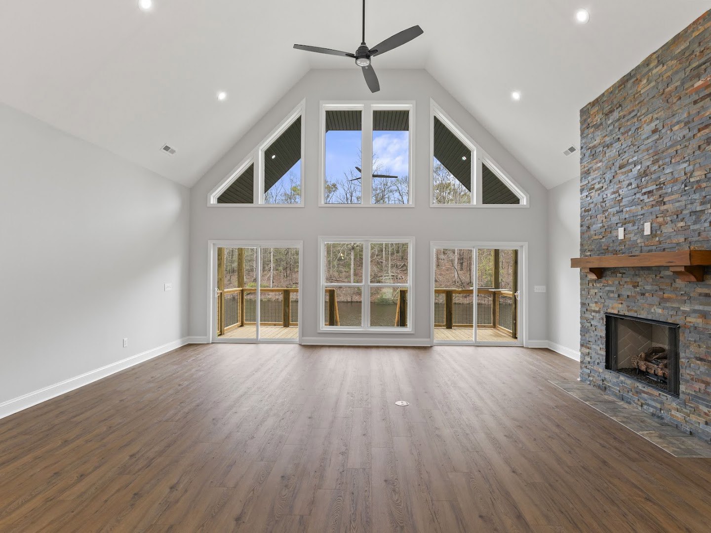 Spacious living room with hardwood floors, white walls, ceiling fan with light, glass-door fireplace, large windows overlooking lake, blue sky, and trees