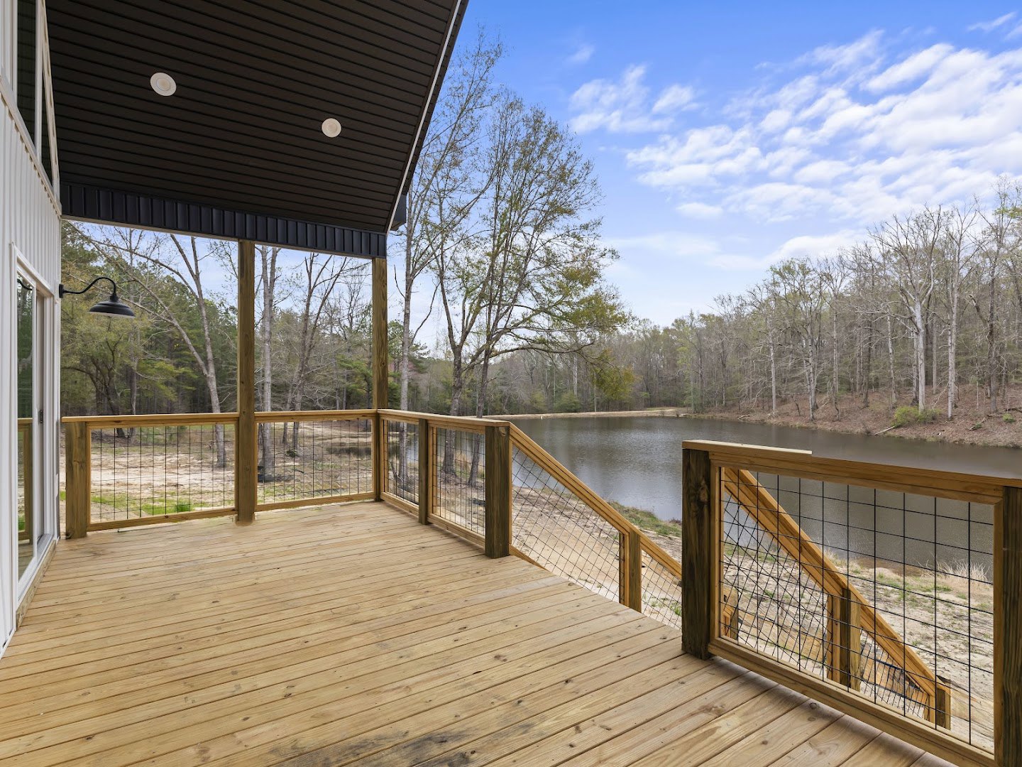 Wooden deck with wire mesh railing, black roof with string lights, overlooking a lake surrounded by trees under a blue sky with clouds