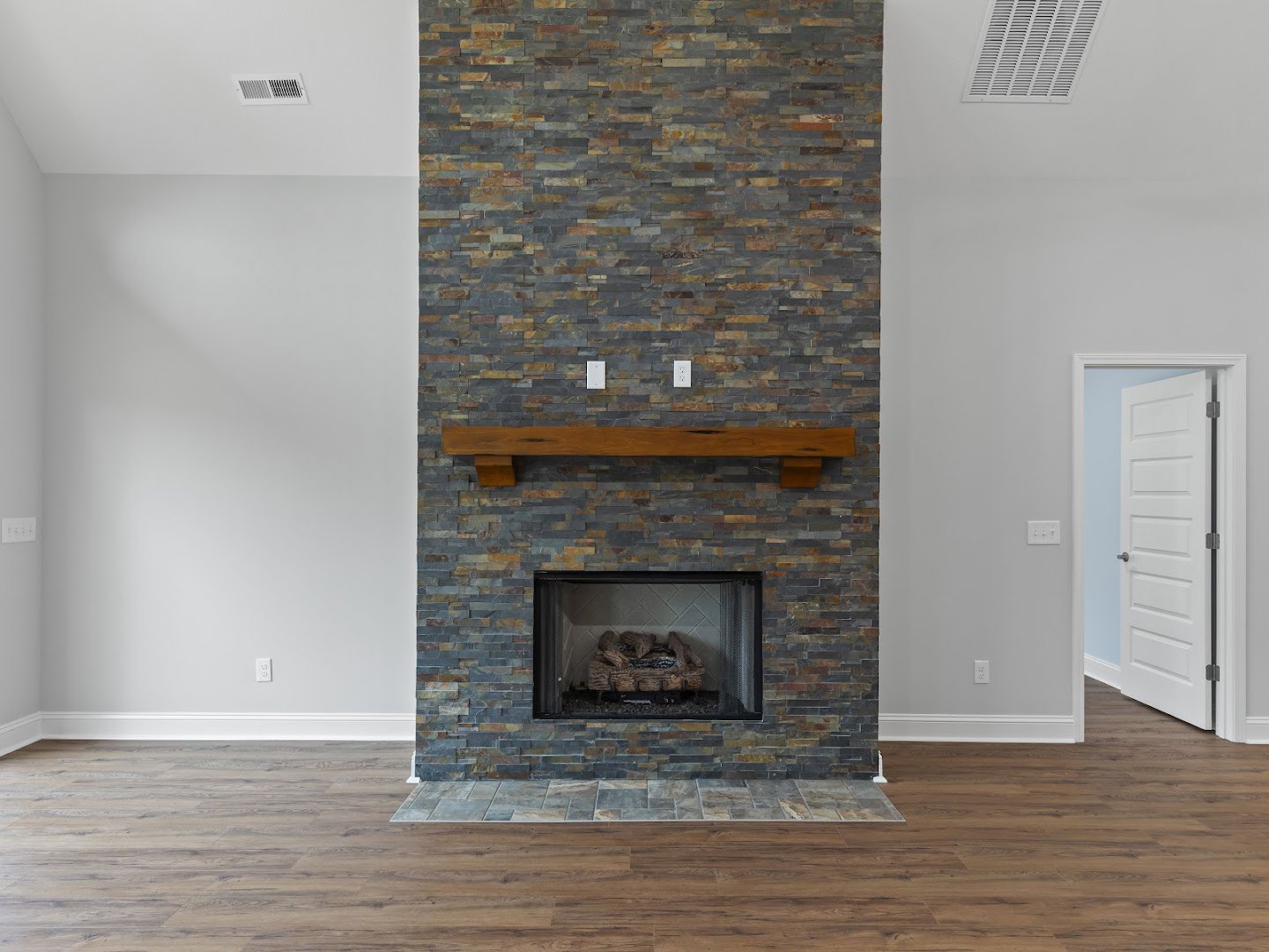 Stone fireplace with wood mantel, stacked logs inside, white door with silver handles, wood bench against stone wall, vent visible near hearth, light-colored flooring