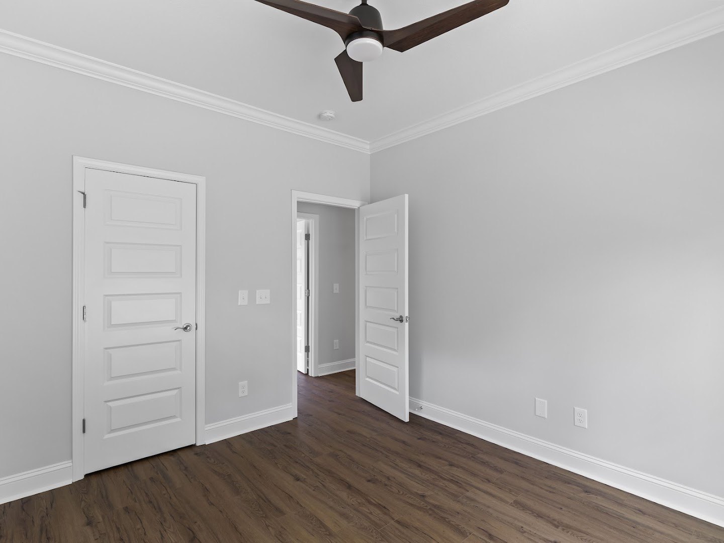 Ceiling fan with light fixture mounted above wood flooring, white walls, and a white door with silver handle and nearby light switch