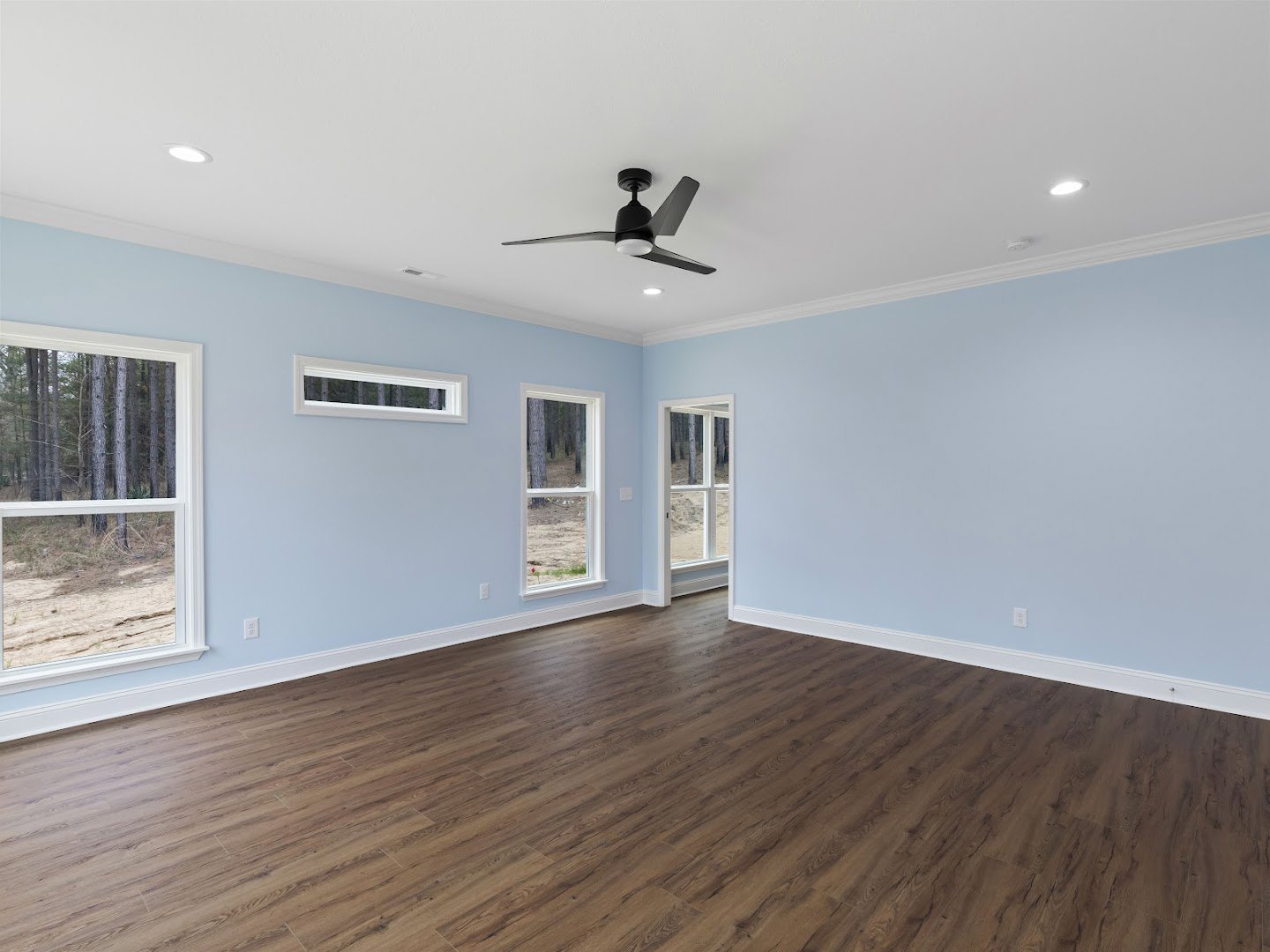 Hardwood floor room with white rectangular windows framing tree views, ceiling fan with light fixture, plaster walls, and wood trim
