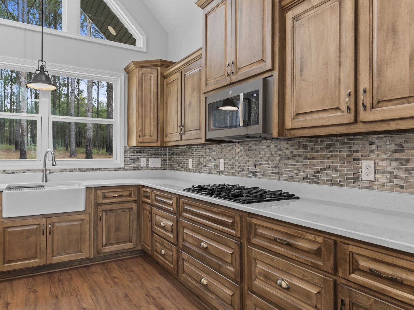 Kitchen with natural wood cabinets, stainless steel microwave above stove, light-colored countertops, modern black metal handles, white tile backsplash, and undermount sink