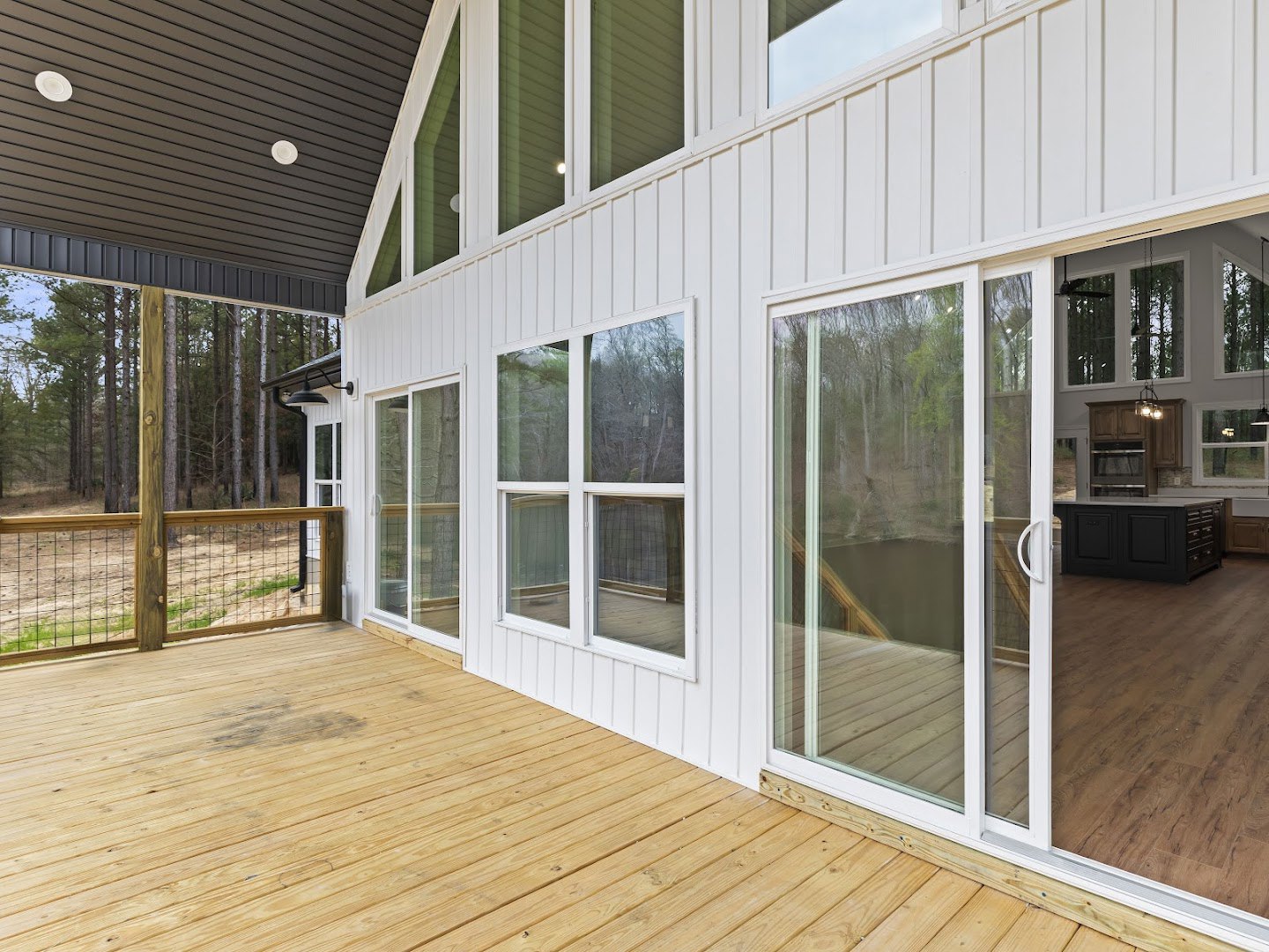 Two-story home with white siding, wooden deck, covered porch, wire mesh fence, large windows, black and white kitchen counters, and trees visible through windows under a clear blue