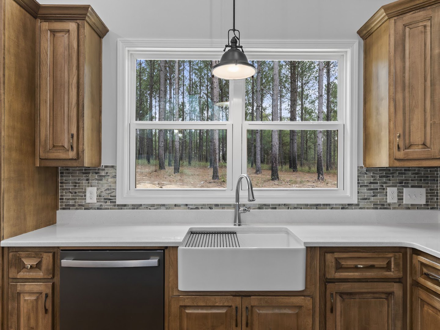 White kitchen sink with metal grate beneath a window overlooking trees, black dishwasher with silver handle, wood cabinetry, black ceiling light fixture, close-up of wood door.