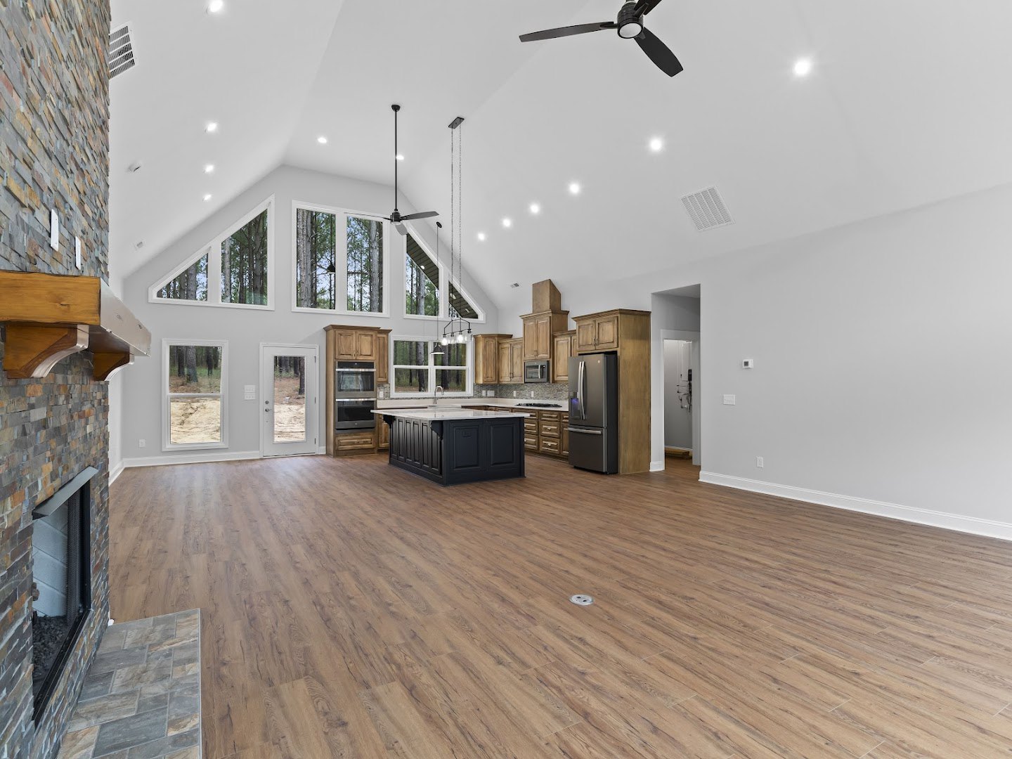 Spacious open floor plan featuring a black kitchen island with white countertops, hardwood floors with central drain, stainless steel refrigerator, exposed brick wall with window