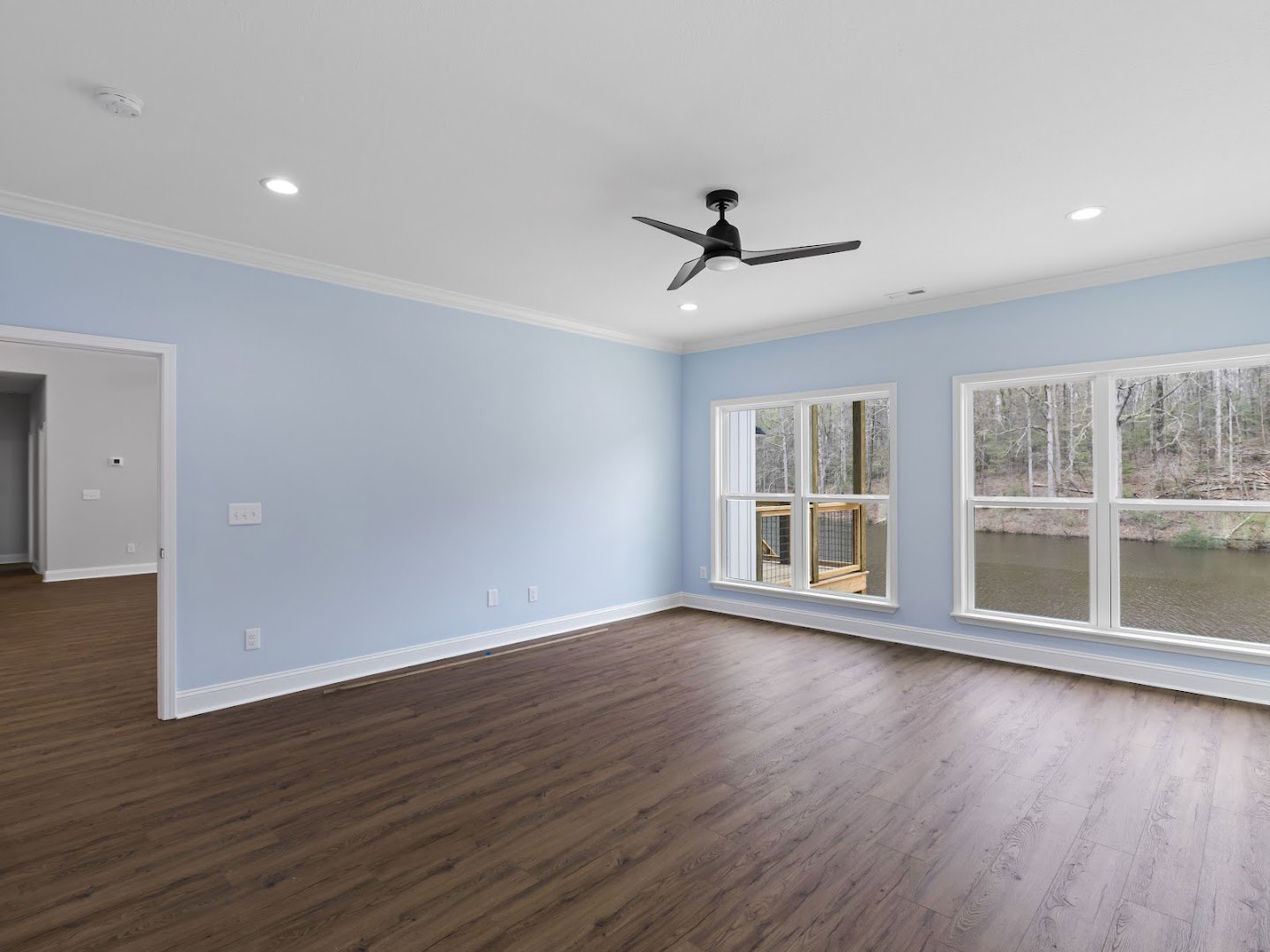Hardwood floor room with ceiling fan, white walls, multiple windows showing river and trees, window ledge with wooden box, white door and light switch