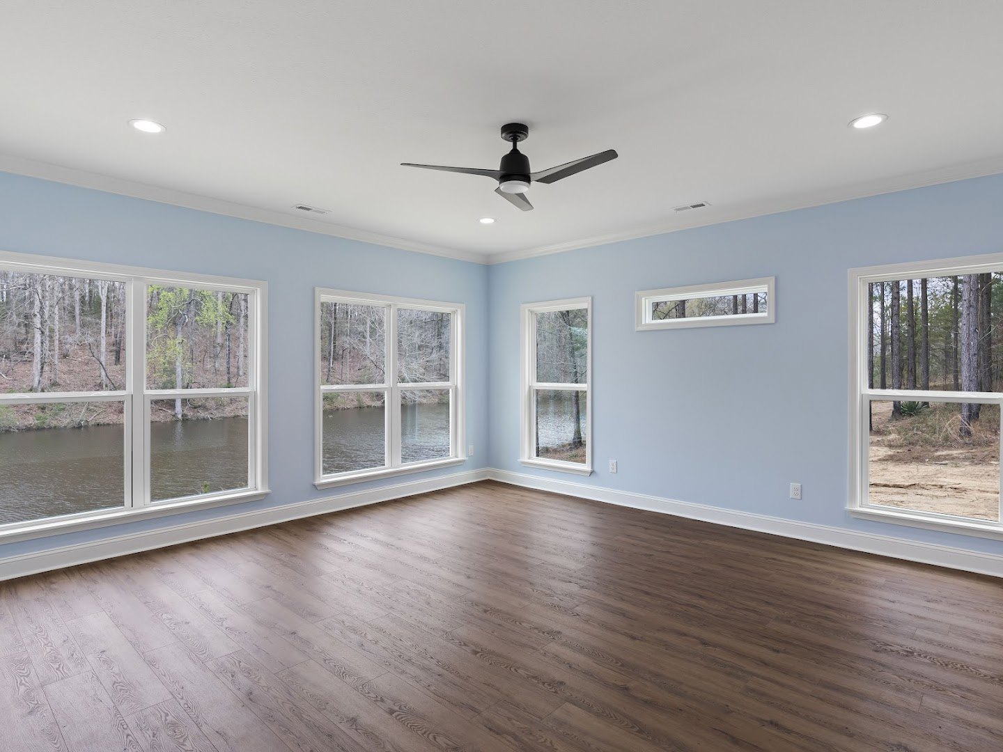 Living room with wood flooring, ceiling fan with light, large windows overlooking trees and river.