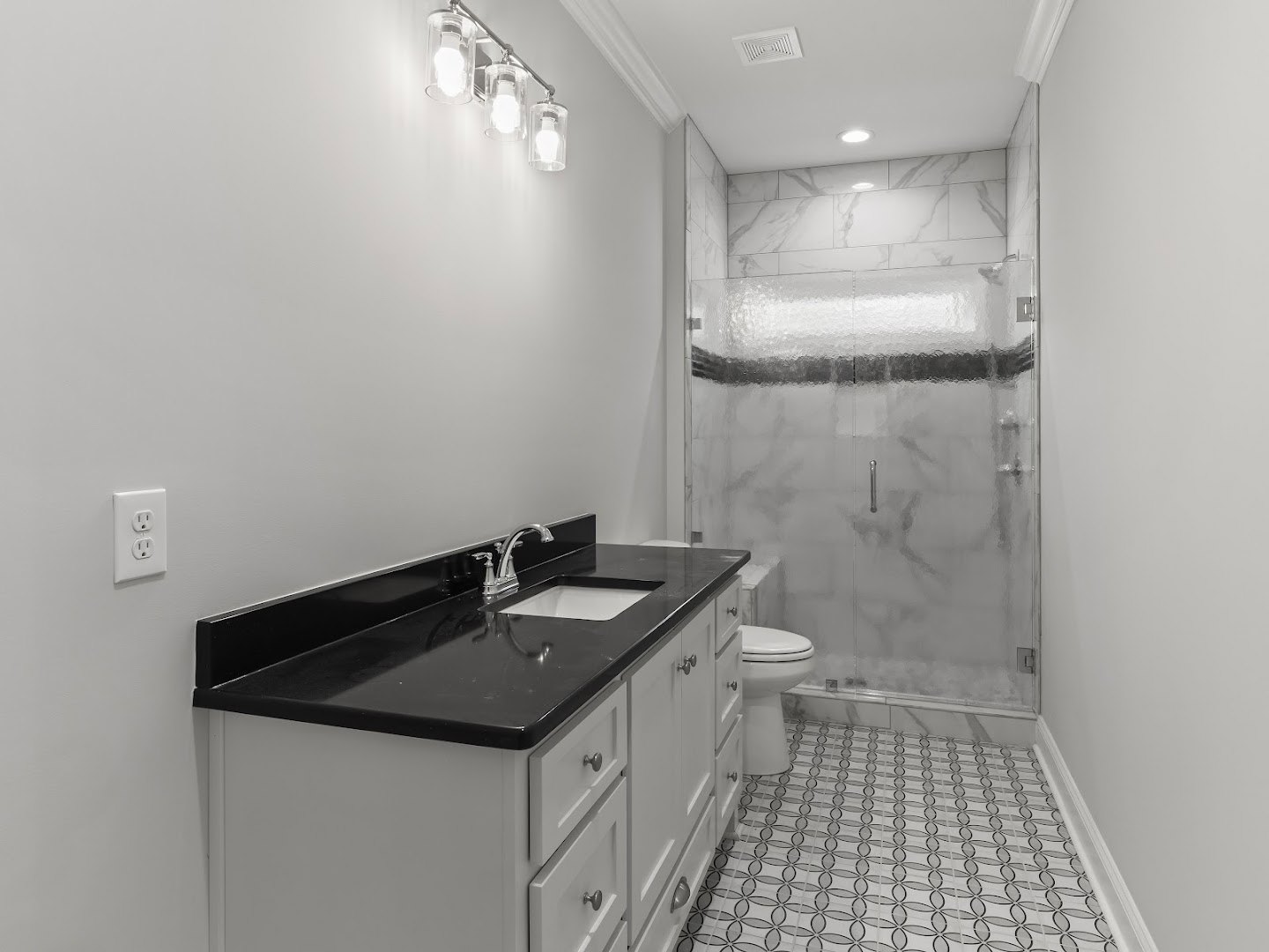 Bathroom featuring a black countertop atop white cabinets, patterned black and white tile floor, glass shower enclosure, ceiling vent, and three-bulb light fixture.
