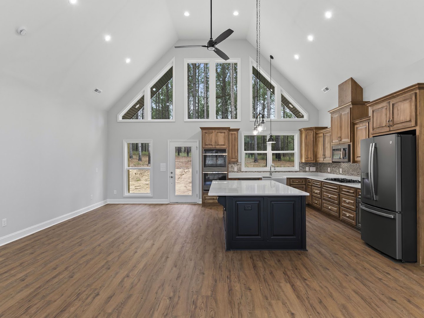 Spacious kitchen featuring a black island with white countertops, hardwood flooring, stainless steel refrigerator, large ceiling fan, white cabinetry, and a window overlooking