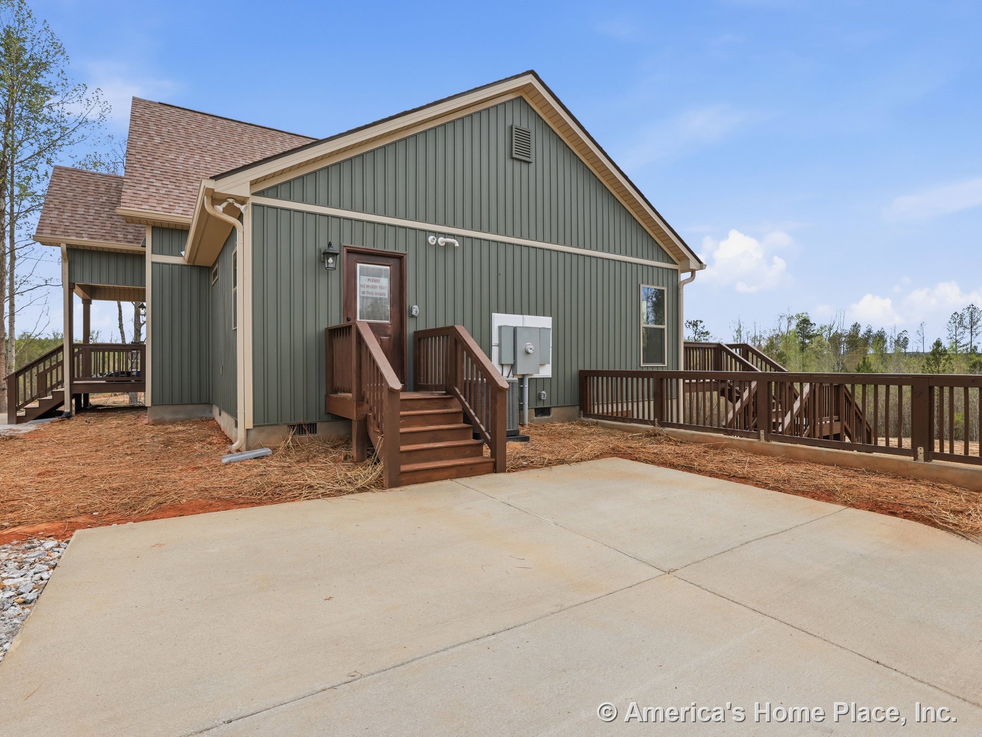 Vertical green siding with gabled shingle roof, covered wooden porch and stairs, wood stair railings, concrete driveway, exterior door with light fixture, window with decorative