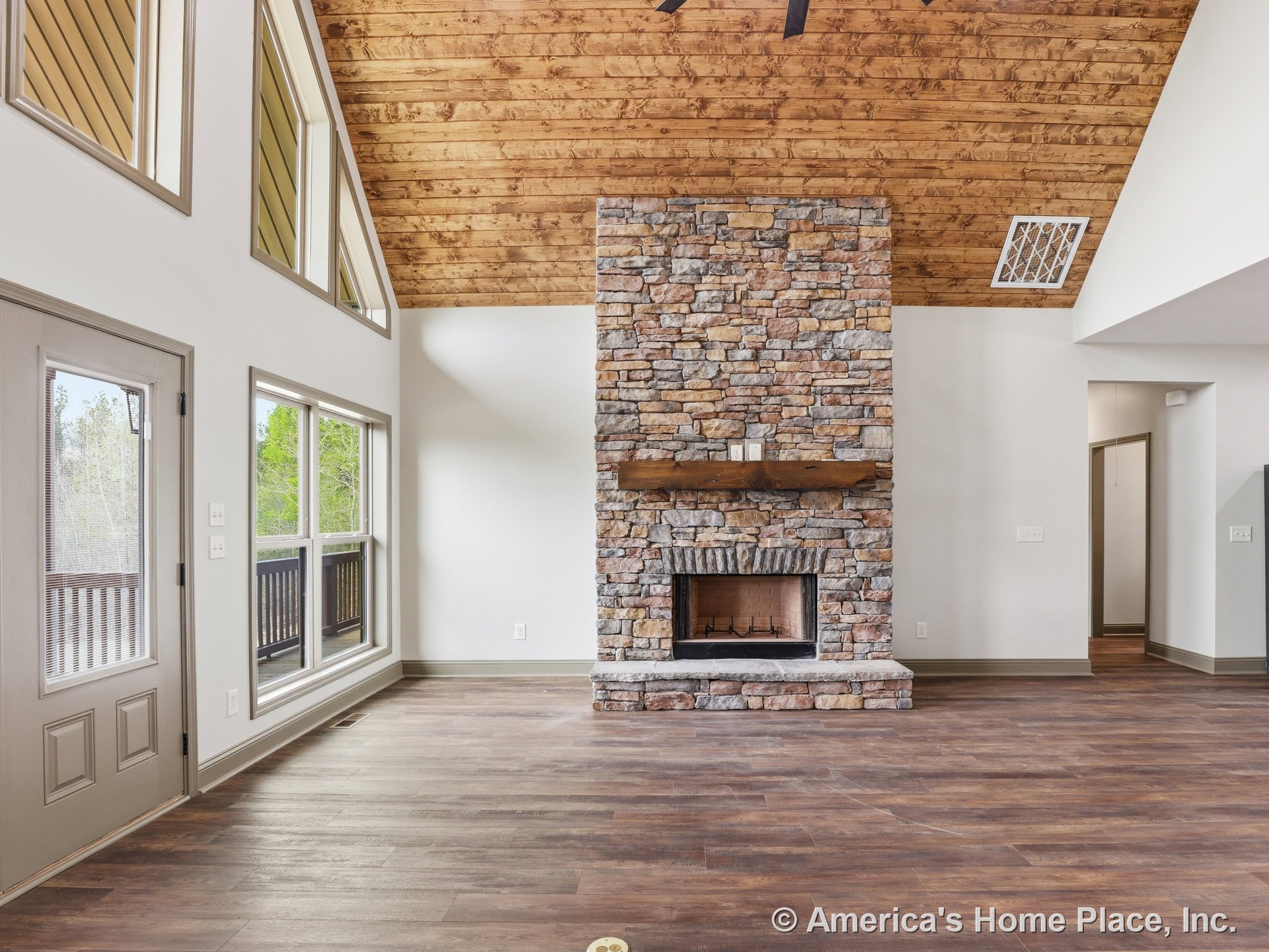 Vaulted wood ceiling and stone fireplace with wood mantel, large windows along one wall, wood-look flooring, trimmed entry door, and abundant natural light.