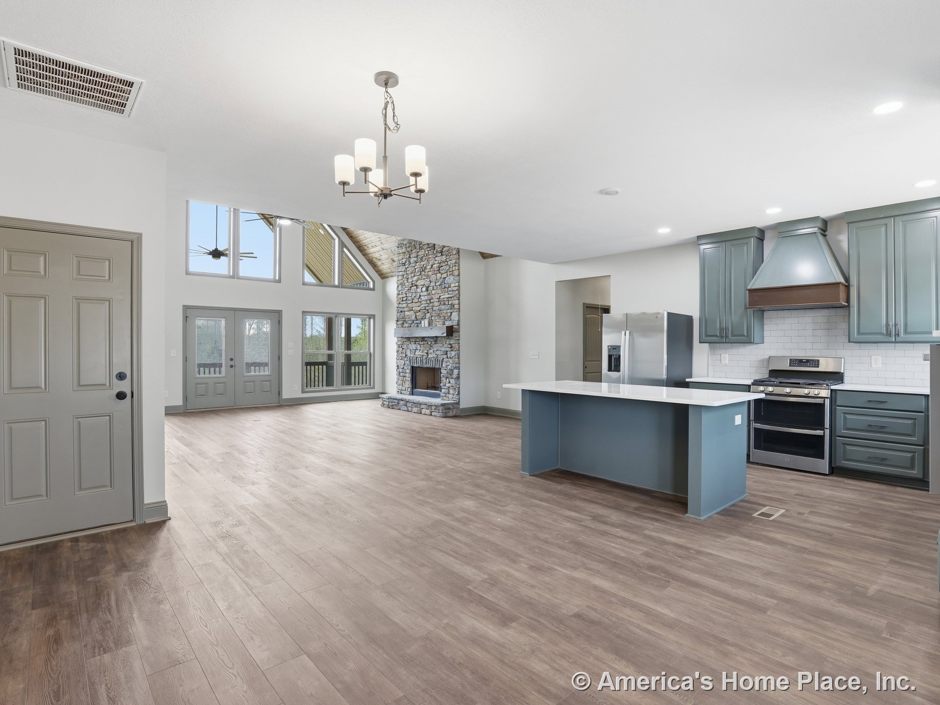 Open-concept living area with stone fireplace and hearth, vaulted ceiling with exposed beams, expansive window wall and glass doors, modern kitchen featuring blue cabinetry and