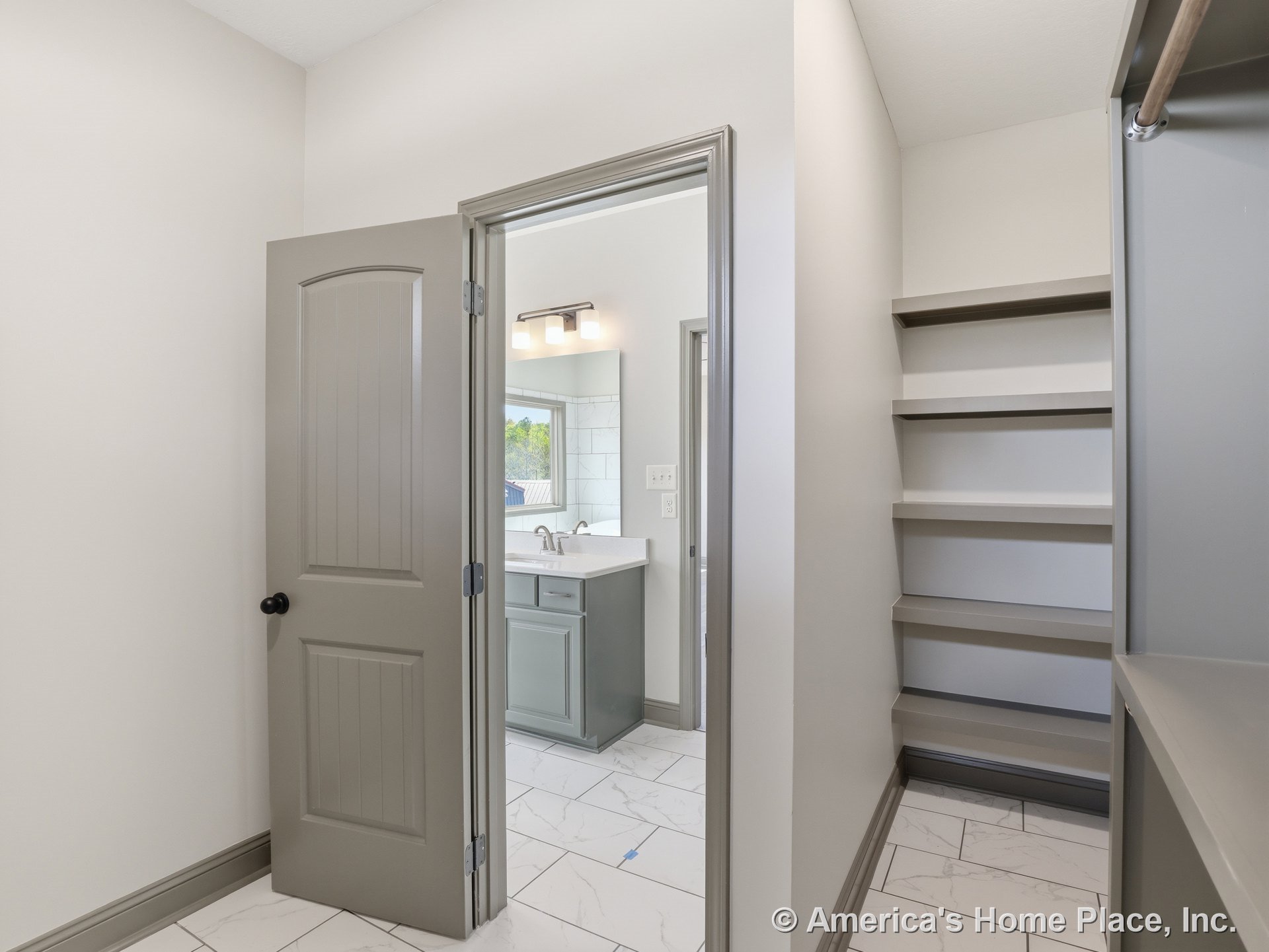 Walk-in closet featuring built-in shelving and cabinetry, connecting to a bathroom with continuous tile flooring, gray paneled door, white vanity with integrated sink, overhead