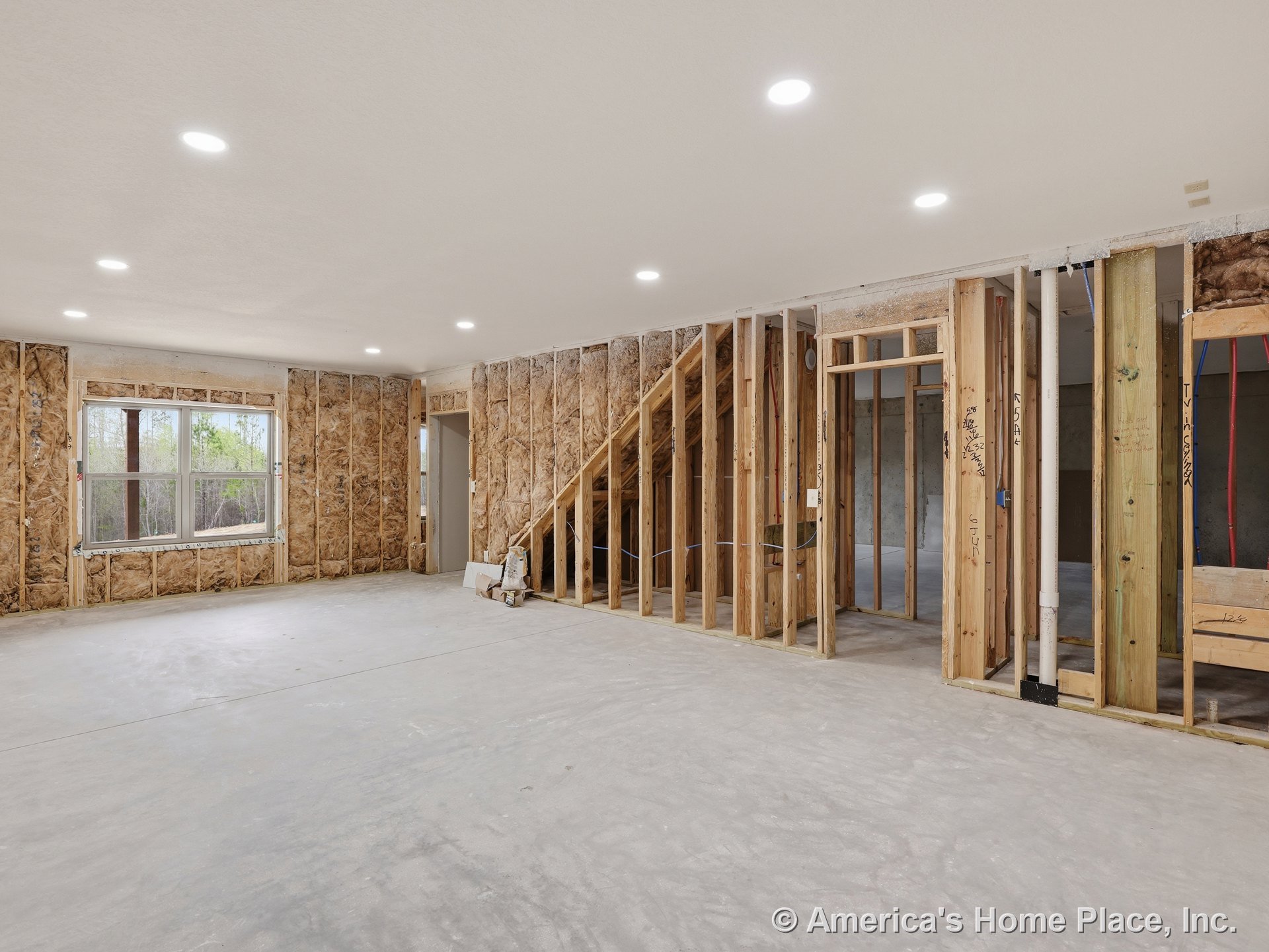 Unfinished basement with exposed wall framing, fiberglass insulation, concrete floor, recessed ceiling lights, large double window, and unfinished staircase framing; visible