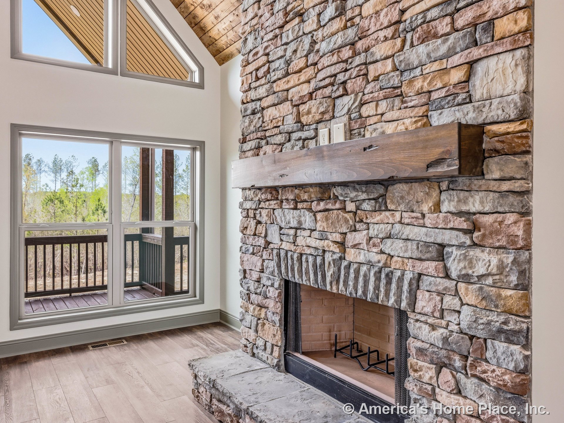 Stacked stone fireplace with wood mantel shelf, vaulted ceiling finished in wood, expansive picture windows overlooking covered porch, light wood flooring, abundant natural light.