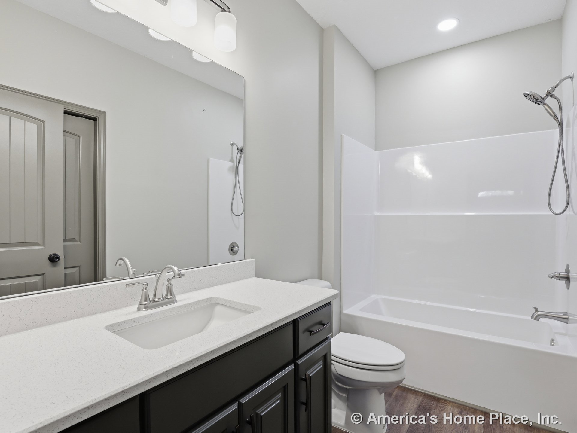 Built-in bathtub with shower fixture beside quartz countertop vanity with dark cabinetry, large wall mirror, recessed ceiling lighting, wood-look flooring, and adjacent sink