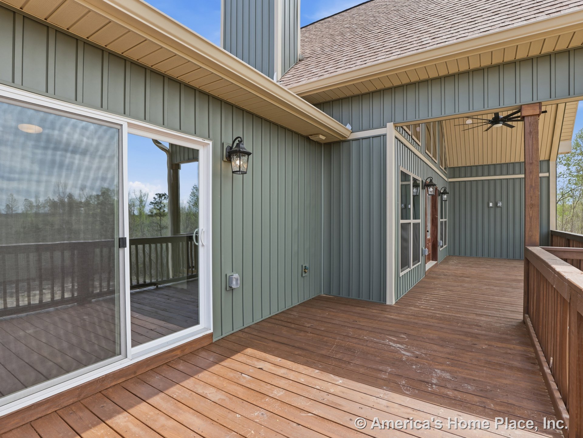 Covered wooden deck with vertical panel siding, multiple windows, sliding glass door, wood plank flooring, outdoor wall lanterns, large ceiling fan, porch ceiling, trim, railing