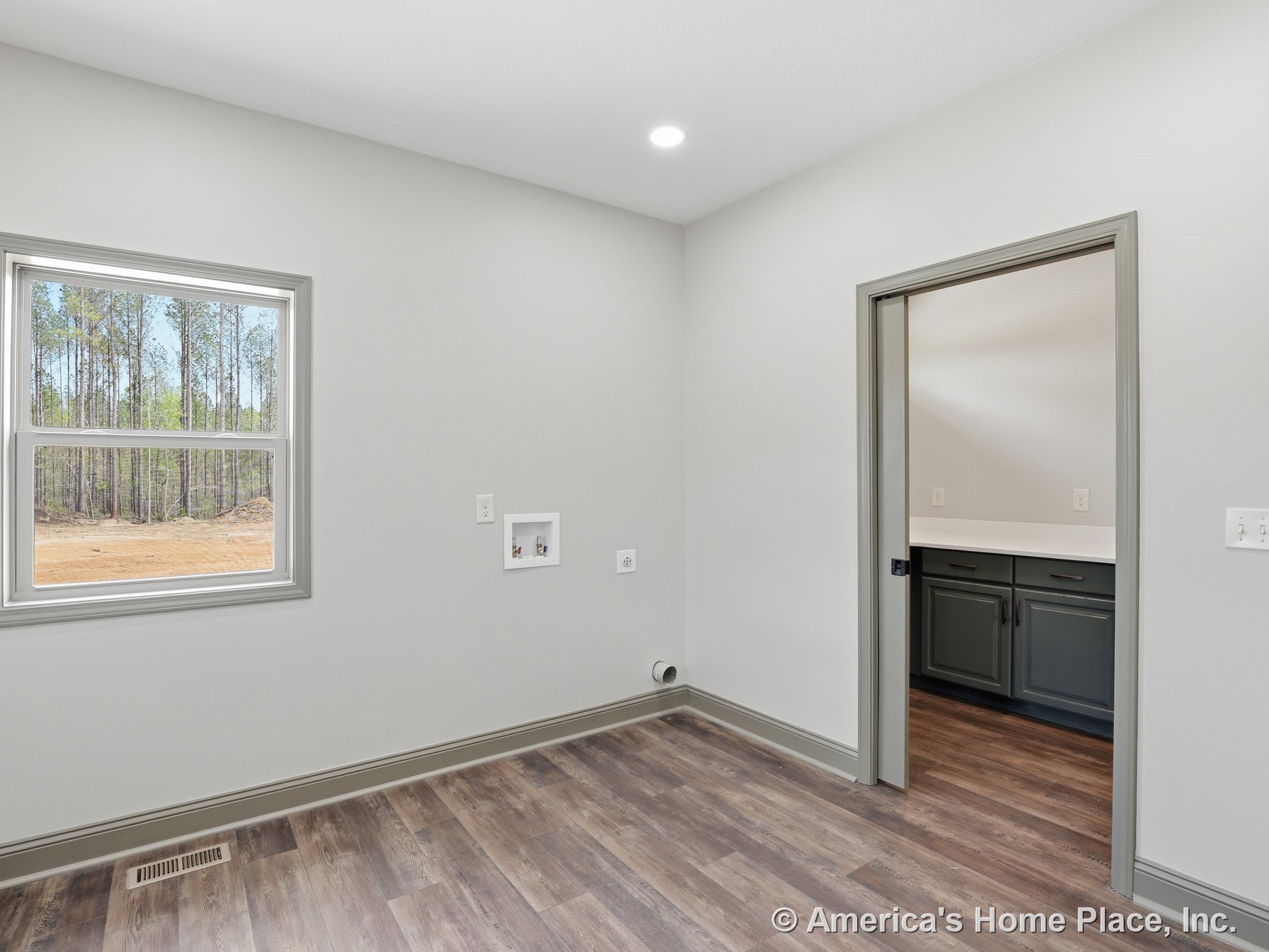 Laundry room with built-in utility hookups on wall, large double-pane window framed by gray trim and baseboards, wood-look vinyl flooring, recessed ceiling light, and adjacent