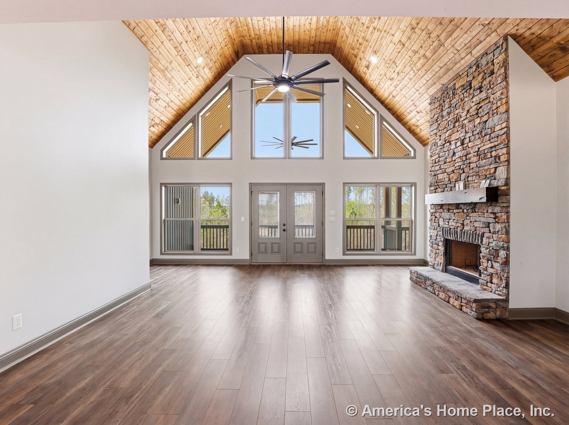 Vaulted wood ceiling with modern ceiling fan, wide plank wood flooring, large triangular windows, stone fireplace with mantel, double doors opening to porch, abundant natural