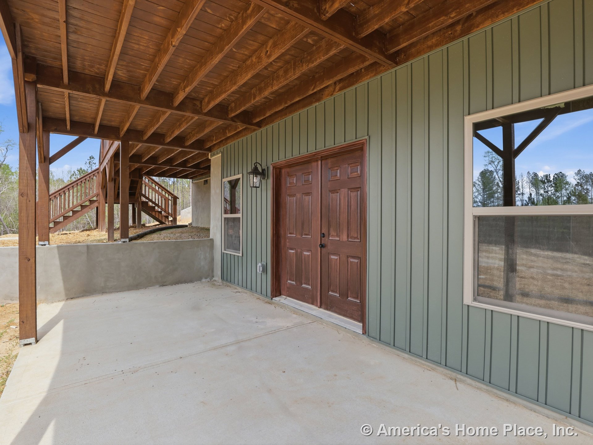 Covered patio with exposed wood beam ceiling, vertical green siding, double wood entry door, concrete flooring, outdoor wall lantern, and adjacent wooden staircase.