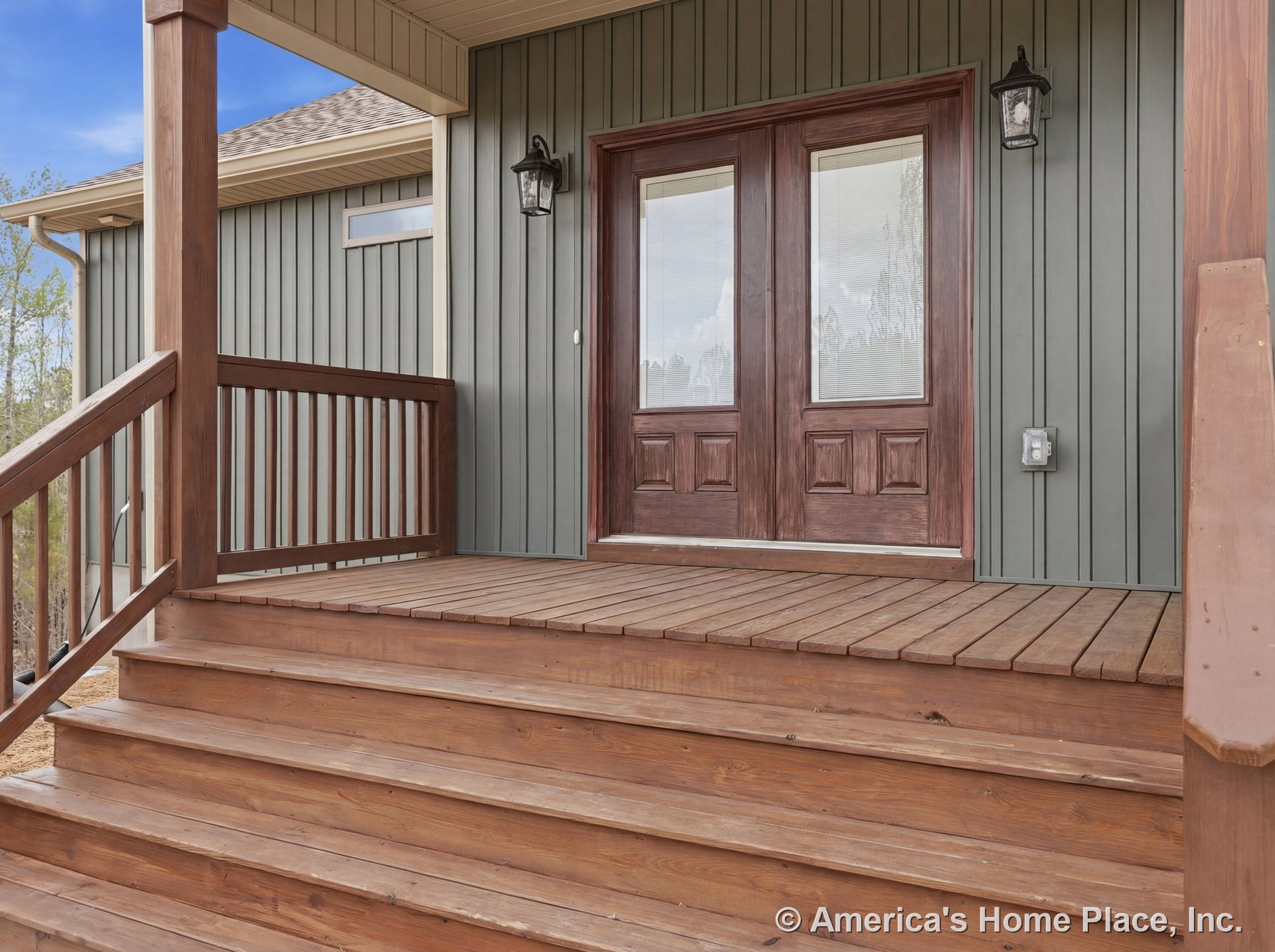 Wood porch steps and railing lead to double glass entry doors set in a vertical siding exterior, with outdoor wall lanterns mounted beside the covered porch ceiling.