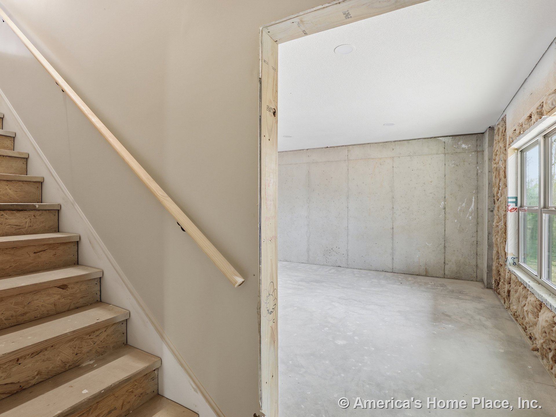 Unfinished basement with exposed concrete walls, large window allowing natural light, plywood stairs, visible insulation, and white ceiling.