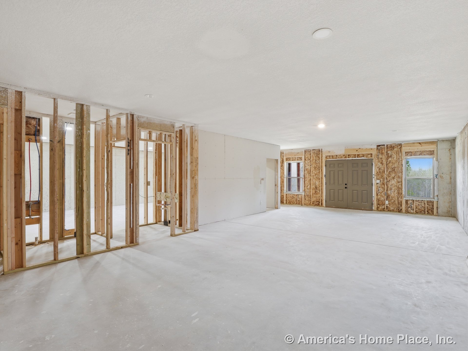 Exposed wood framing and unfinished drywall surround a main room with a double entry door, concrete flooring, two windows, and recessed ceiling lights; no cabinetry or trim