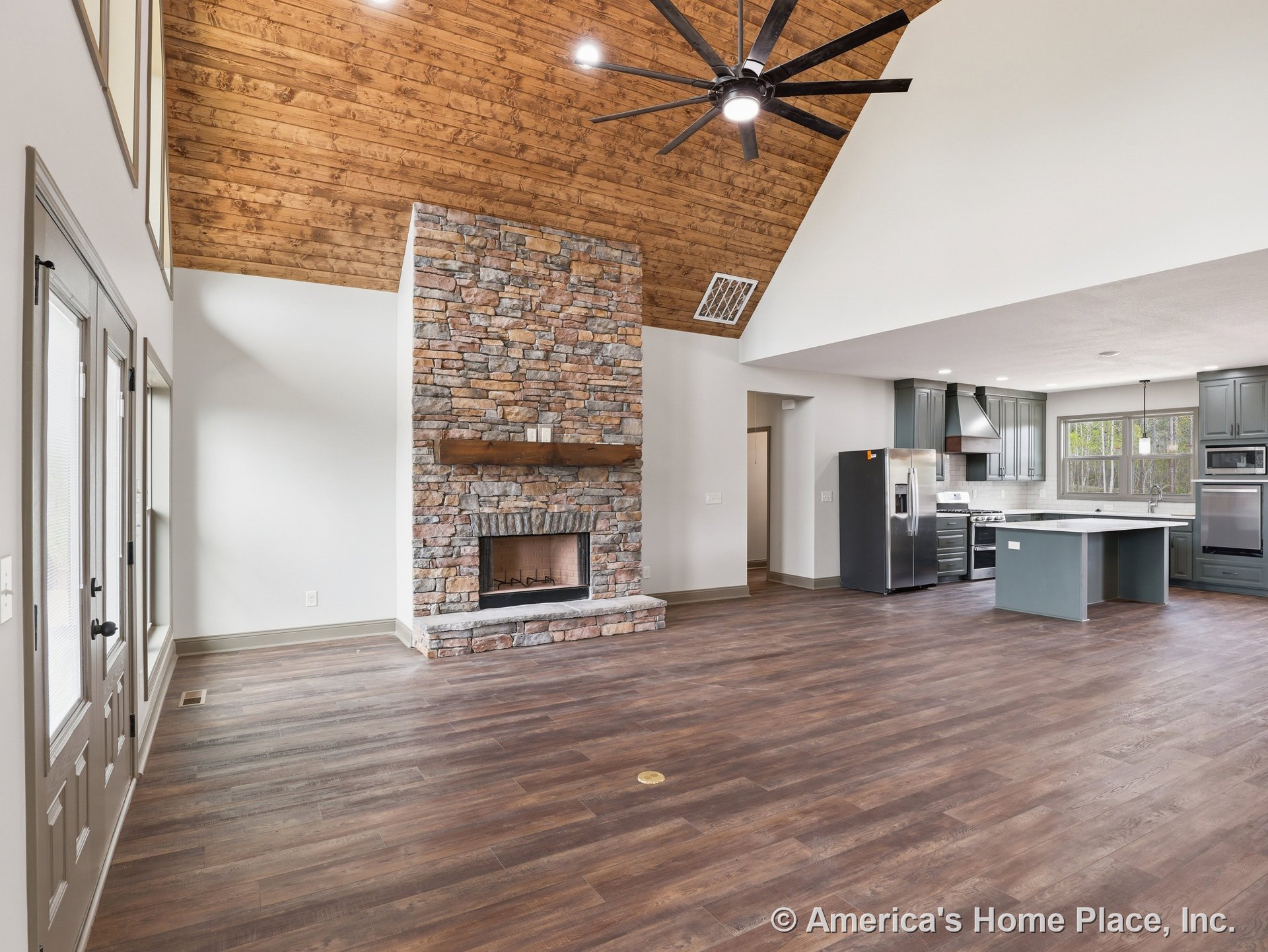 Vaulted wood ceiling and stone fireplace with mantel in open living area, featuring modern kitchen with gray built-in cabinetry, stainless appliances, large windows, hardwood