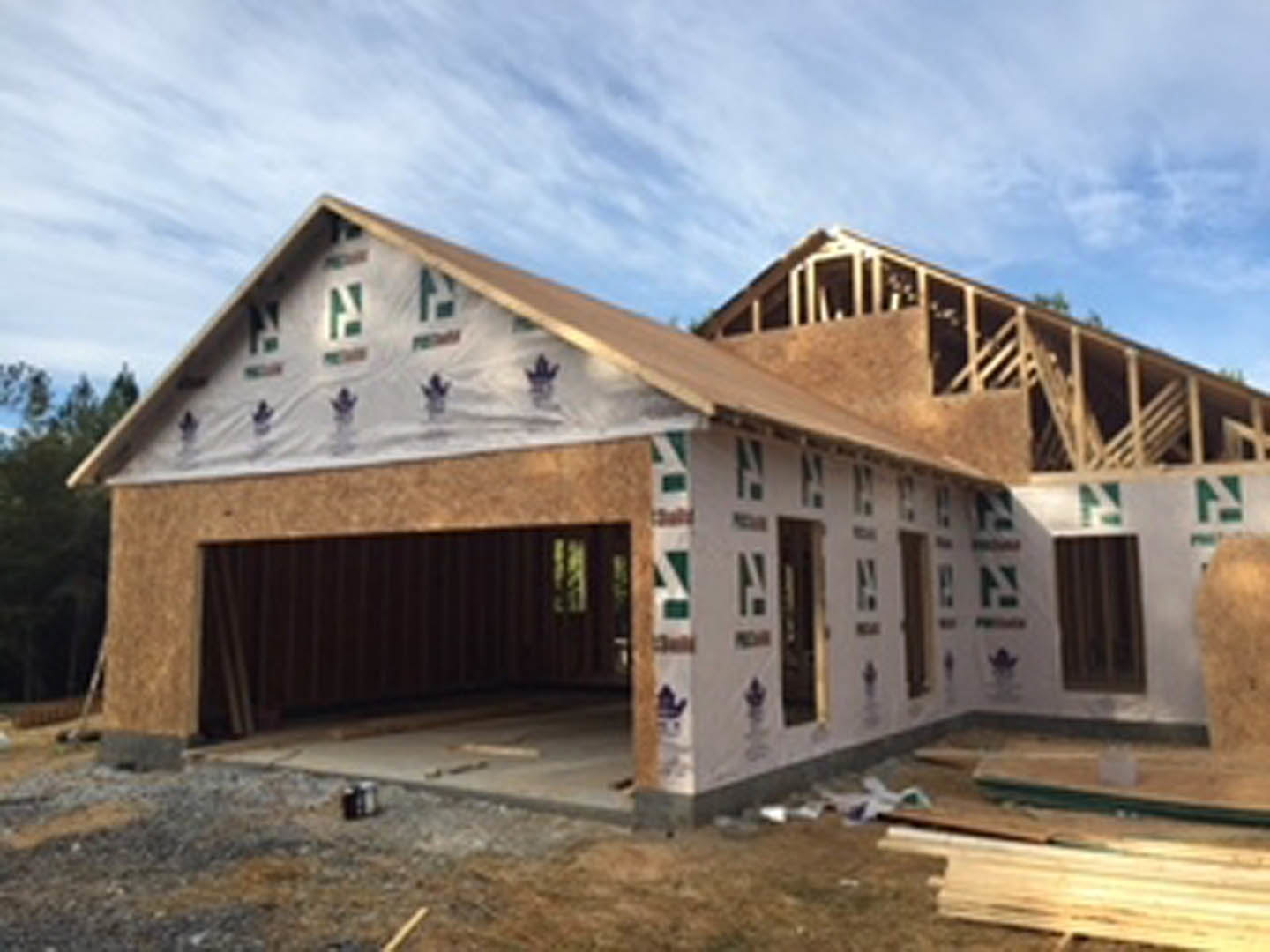 Wood-framed house under construction with exposed beams, partially completed roof, and open exterior walls on a dirt lot under a cloudy sky