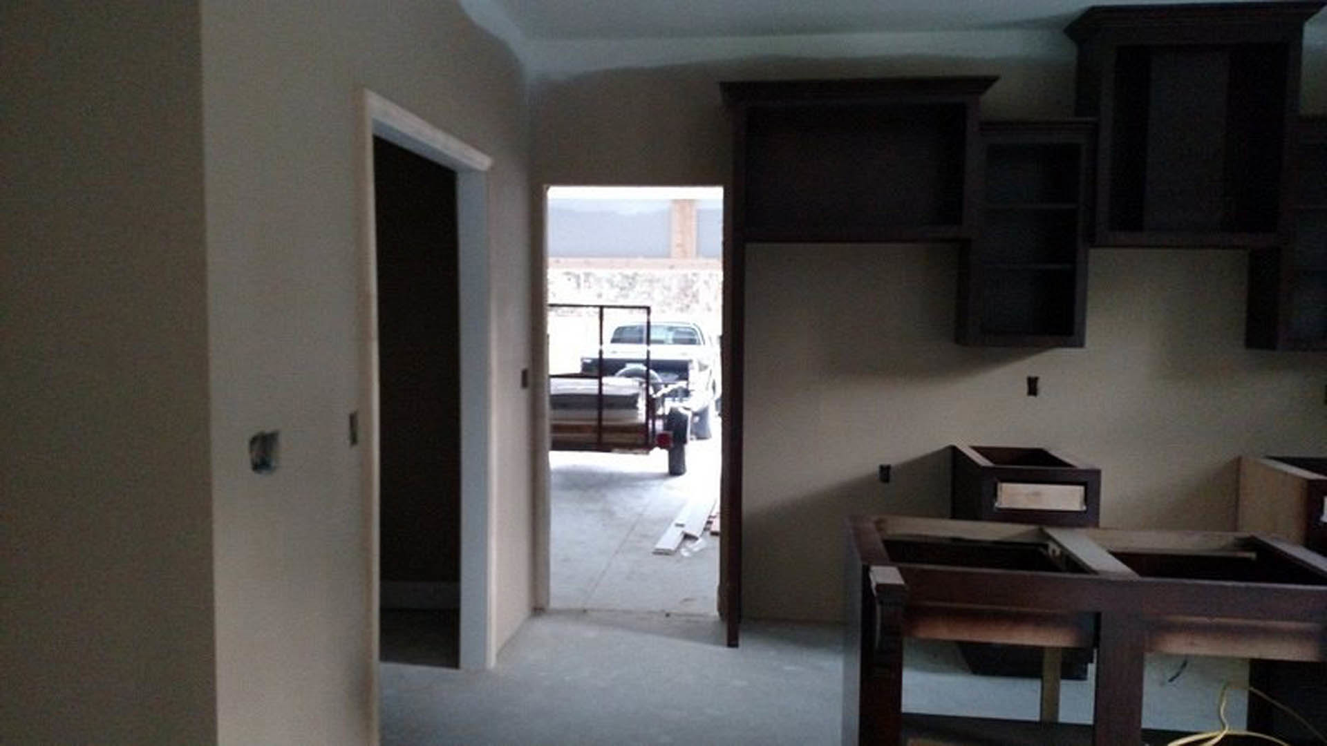 Open door in a residential room reveals parked truck outside; wood table and chair sit on light flooring, white walls and cabinetry visible.