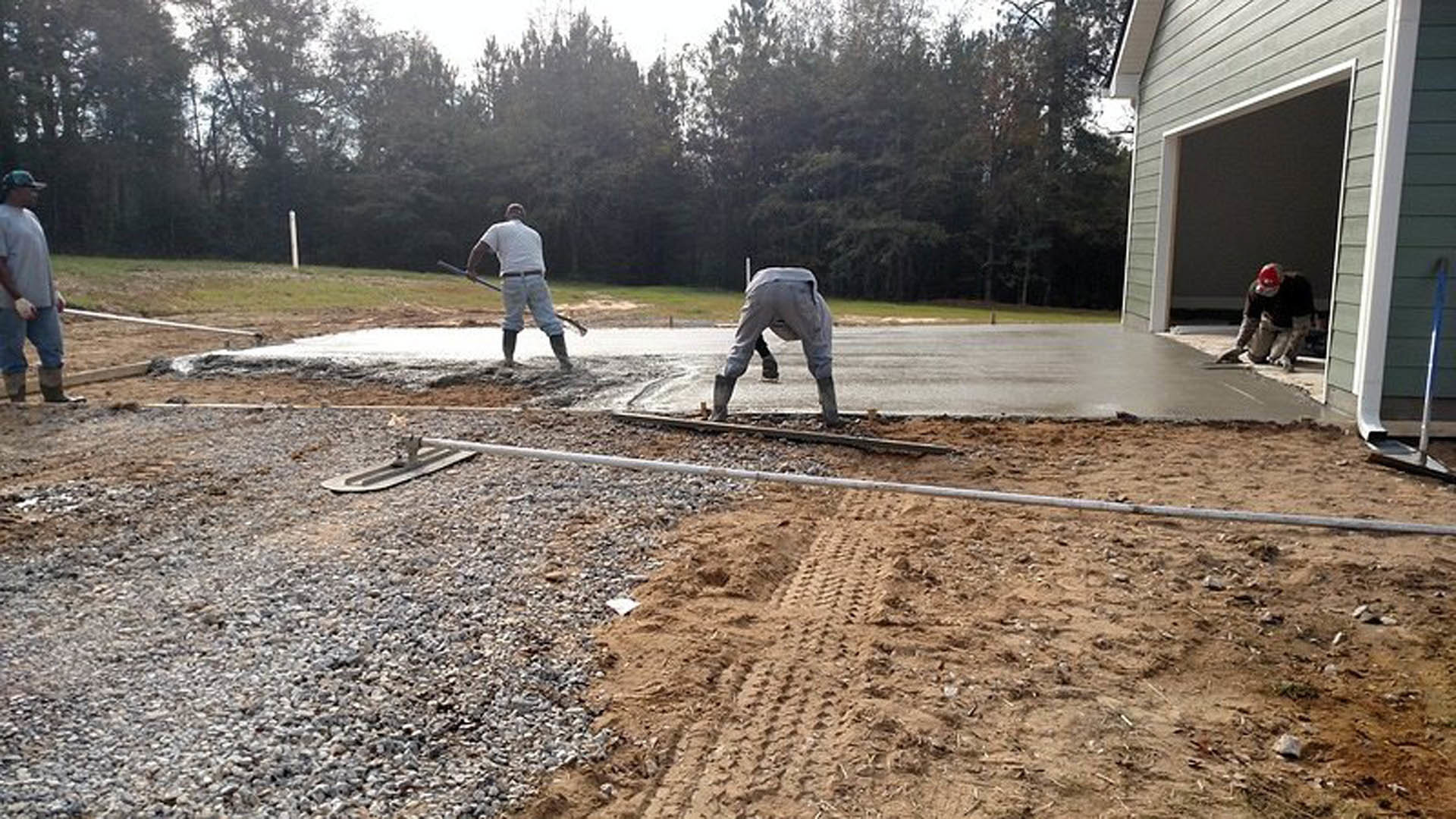 Two men working on a concrete slab foundation outdoors, one in grey pants and boots holding a stick, another wearing a helmet, surrounded by soil, trees, and construction