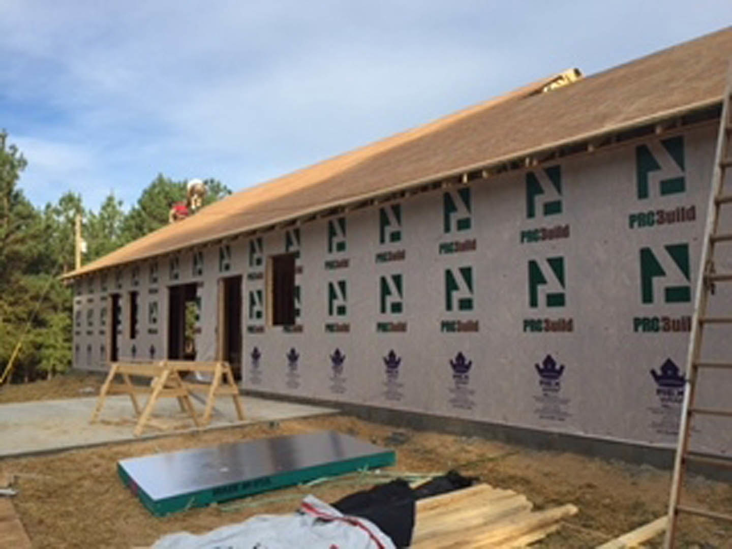 Framed custom home under construction with exposed lumber, workers on site, cloudy sky overhead, trees in background