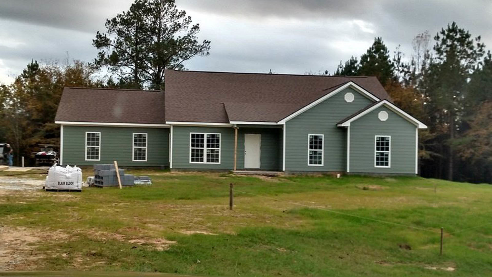 Two-story house with gray siding, white trim, and a white front door, surrounded by a green lawn and bordered by a wooden fence; large windows and cloudy sky in background.