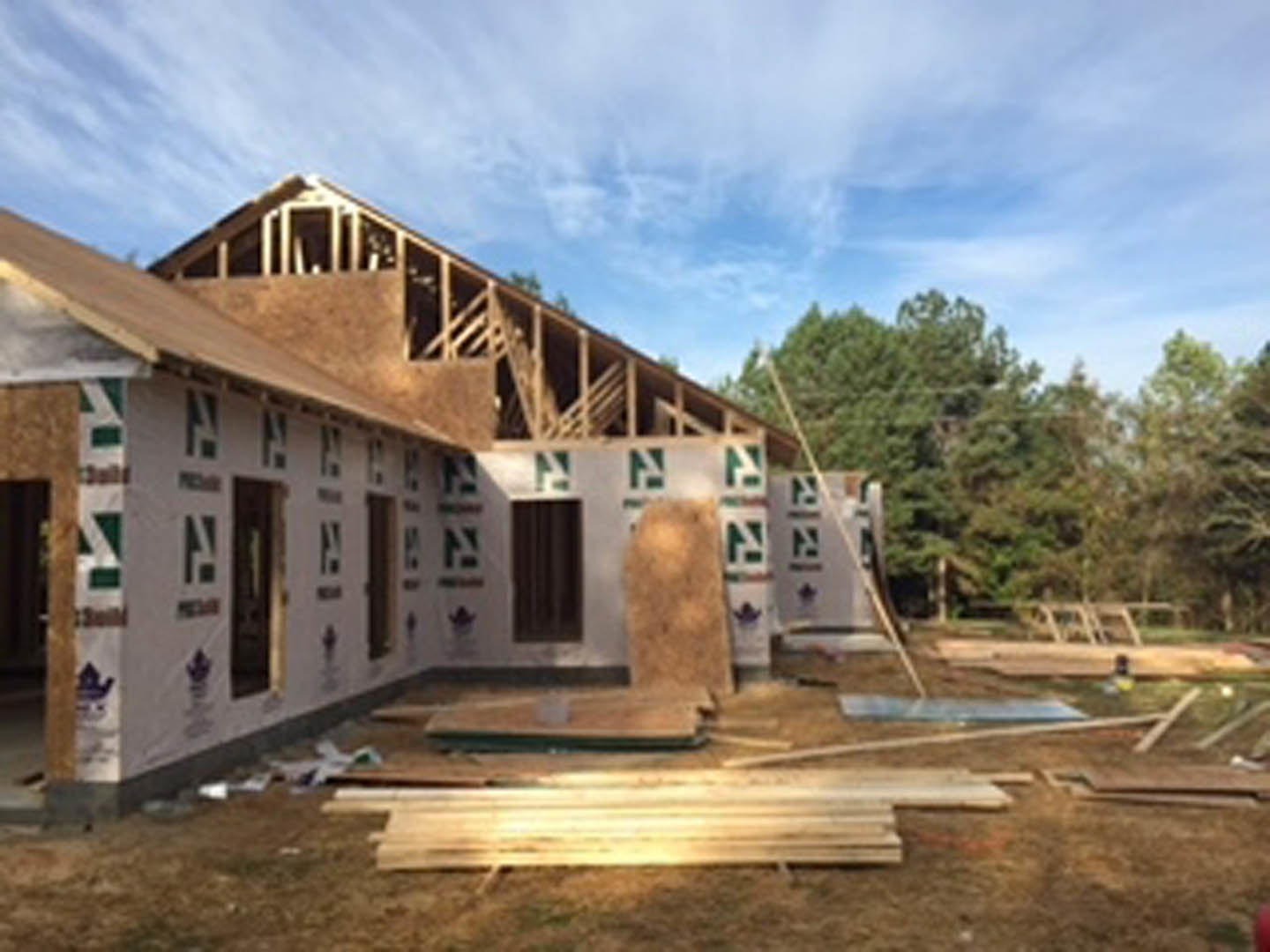 Framed house under construction with stacks of lumber on dirt ground, cloudy sky overhead, trees in background