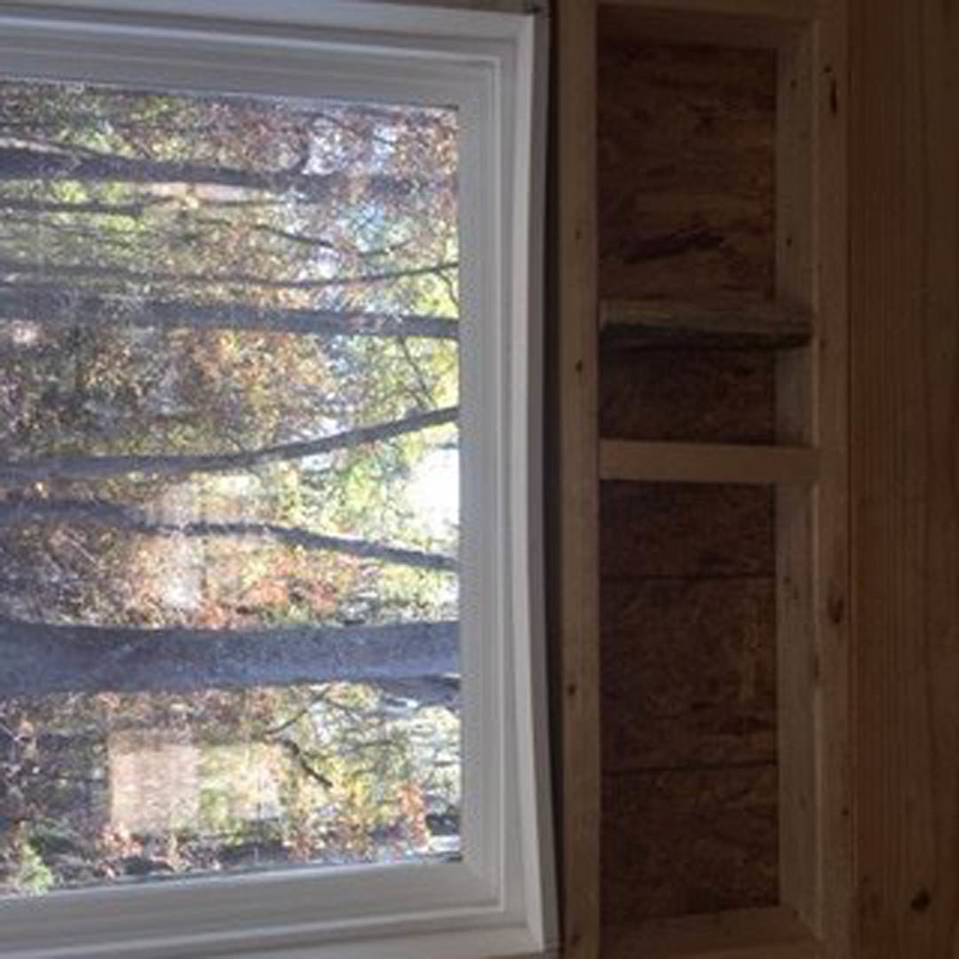 Large window with view of green trees, built-in wooden shelf beneath, wood panel wall, wooden ladder leaning nearby in bright room
