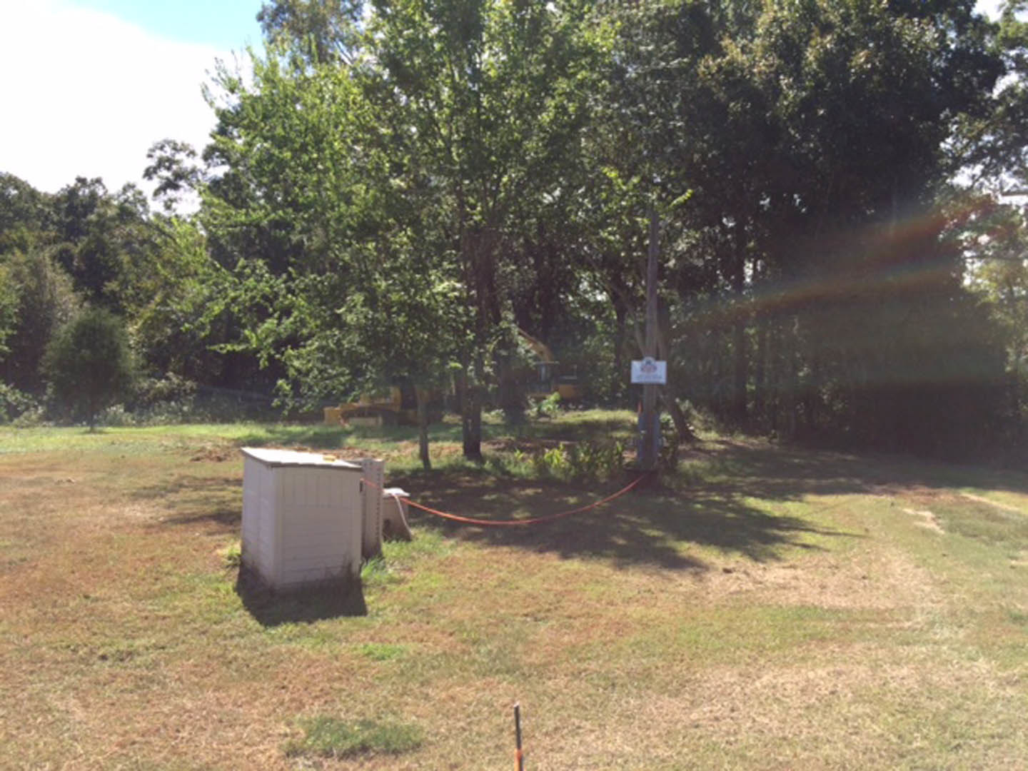 Fenced grassy area with mature trees, white shed, and a white sign featuring red and blue design; rope tied to building and person walking in background.