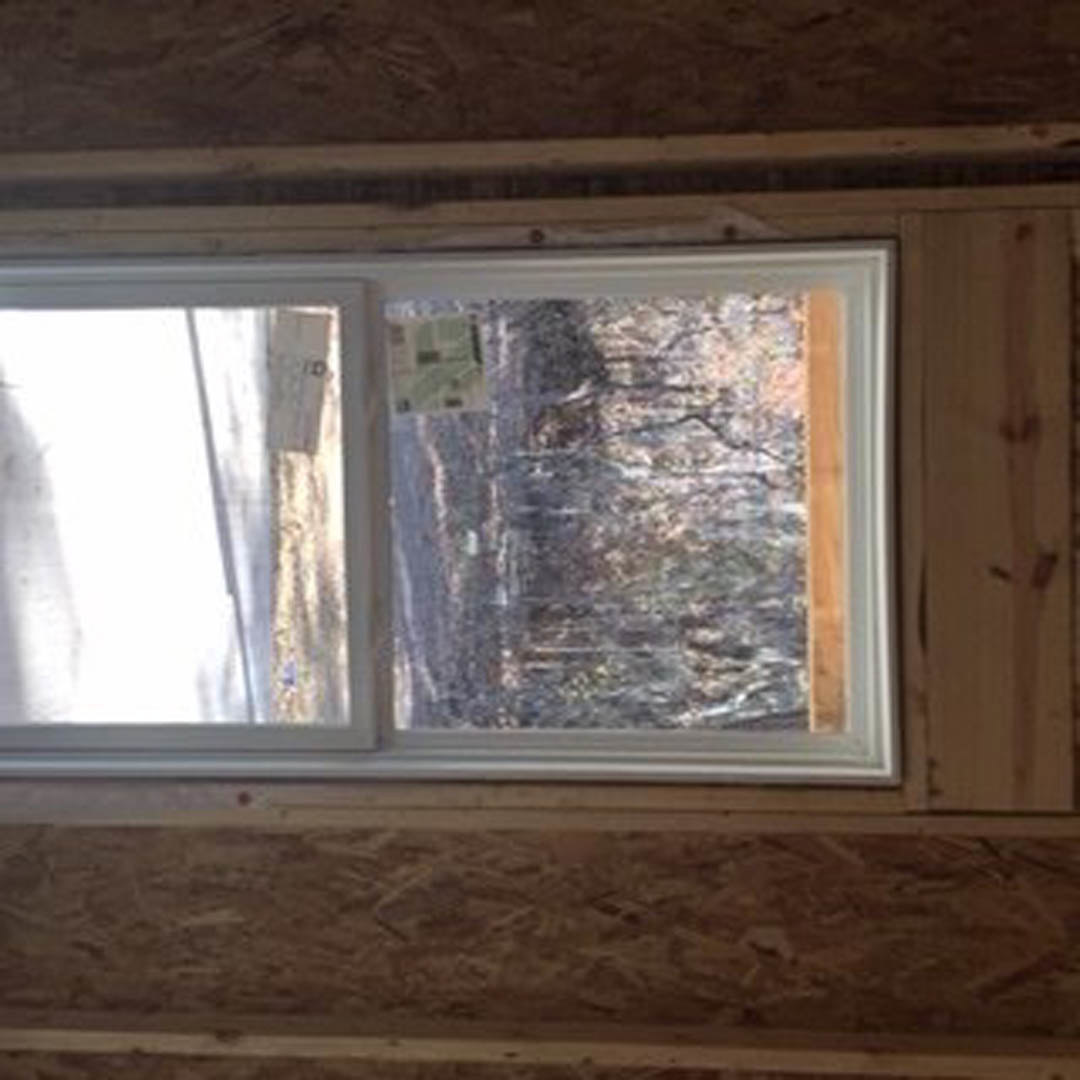 Large window framed in natural wood, overlooking dense forest, with sunlight filtering into a modern room; paper taped to glass and blurred cat visible on windowsill.