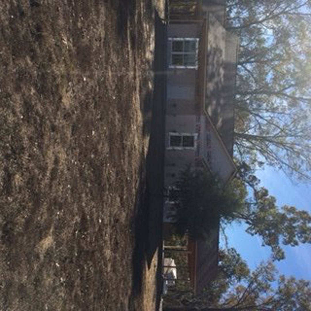 Two-story custom home with light siding, large windows, and gabled roof, surrounded by green yard and mature trees under blue sky