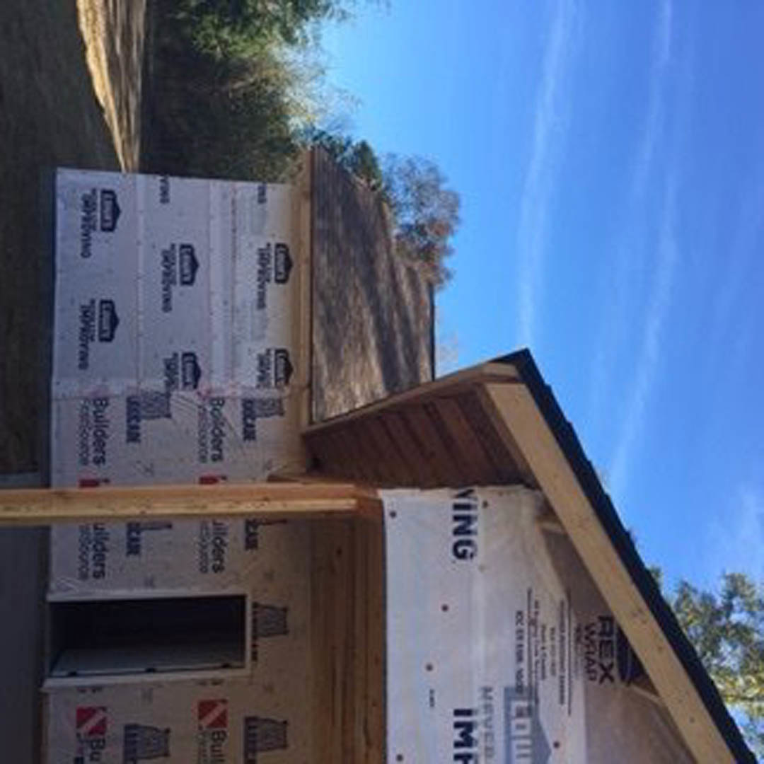 Wood-framed house under construction with exposed beams, partially completed roof, surrounded by trees and cloudy sky