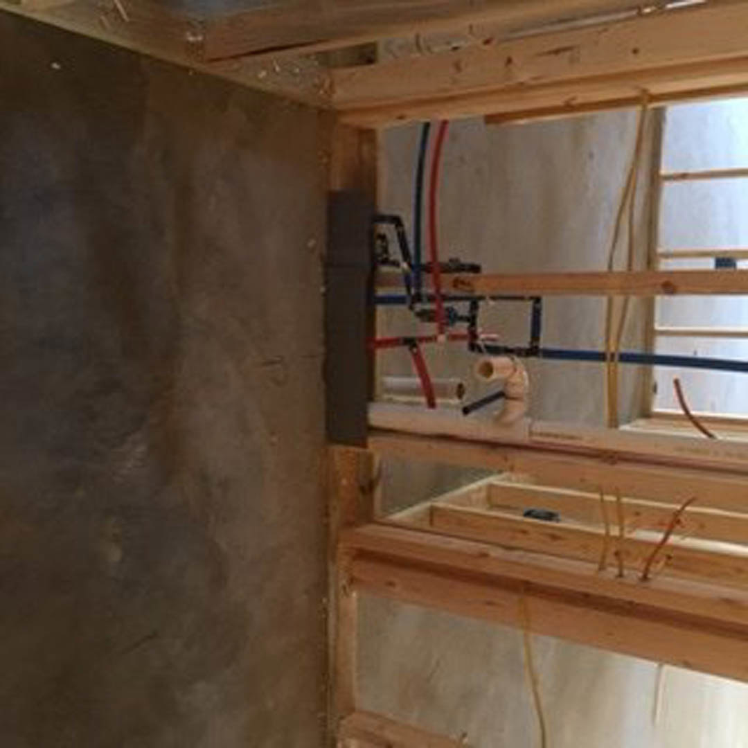 Wood framing with exposed pipes, insulation, and plywood panels in an unfinished room; blurred hand partially visible in foreground