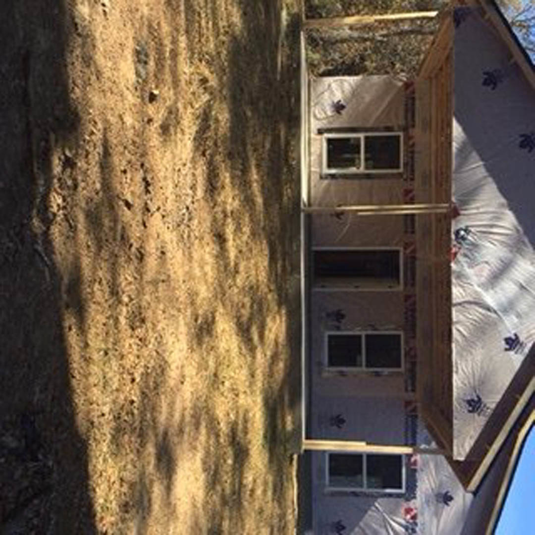 Two-story custom home under construction with exposed framing, large windows, and a patch of bare dirt in the foreground