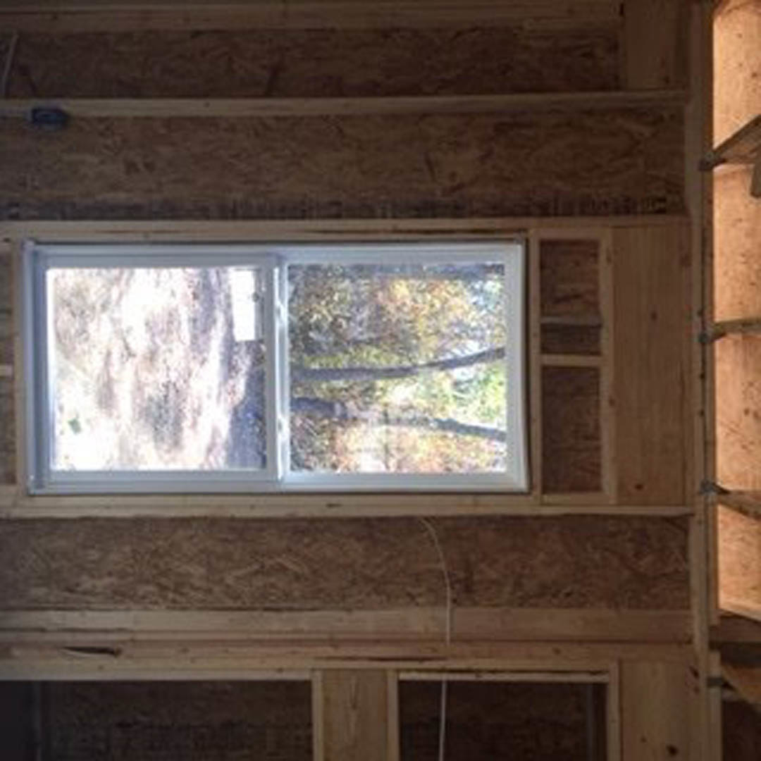 Large wood-framed window in a bright room, offering a view of dense forest through clear glass, surrounded by white walls and natural wood trim