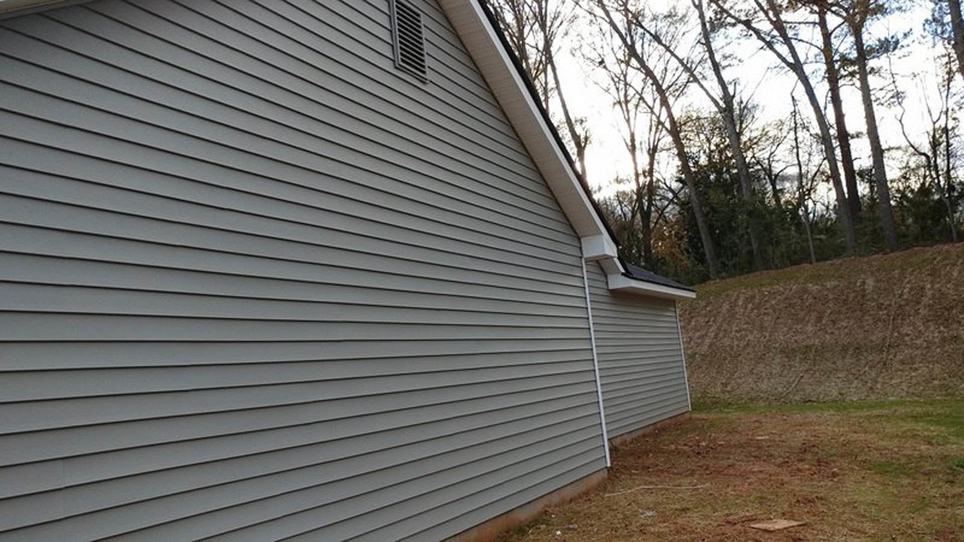 Grey siding and white roof on a house with a grassy lawn, dirt patch, and mature trees in the background; person standing on a hill near the property, close-up of exterior vent