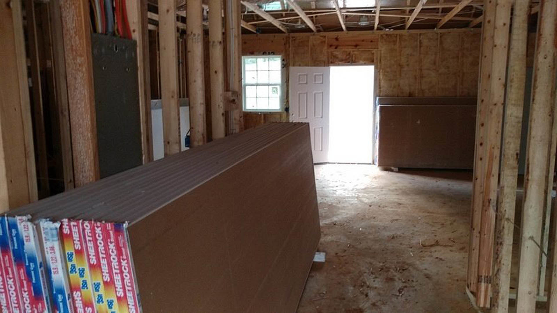 Living room with exposed wood ceiling beams, large rectangular storage box, white-framed window, stacked sheets against wall, white door illuminated by bright light, boxes arranged