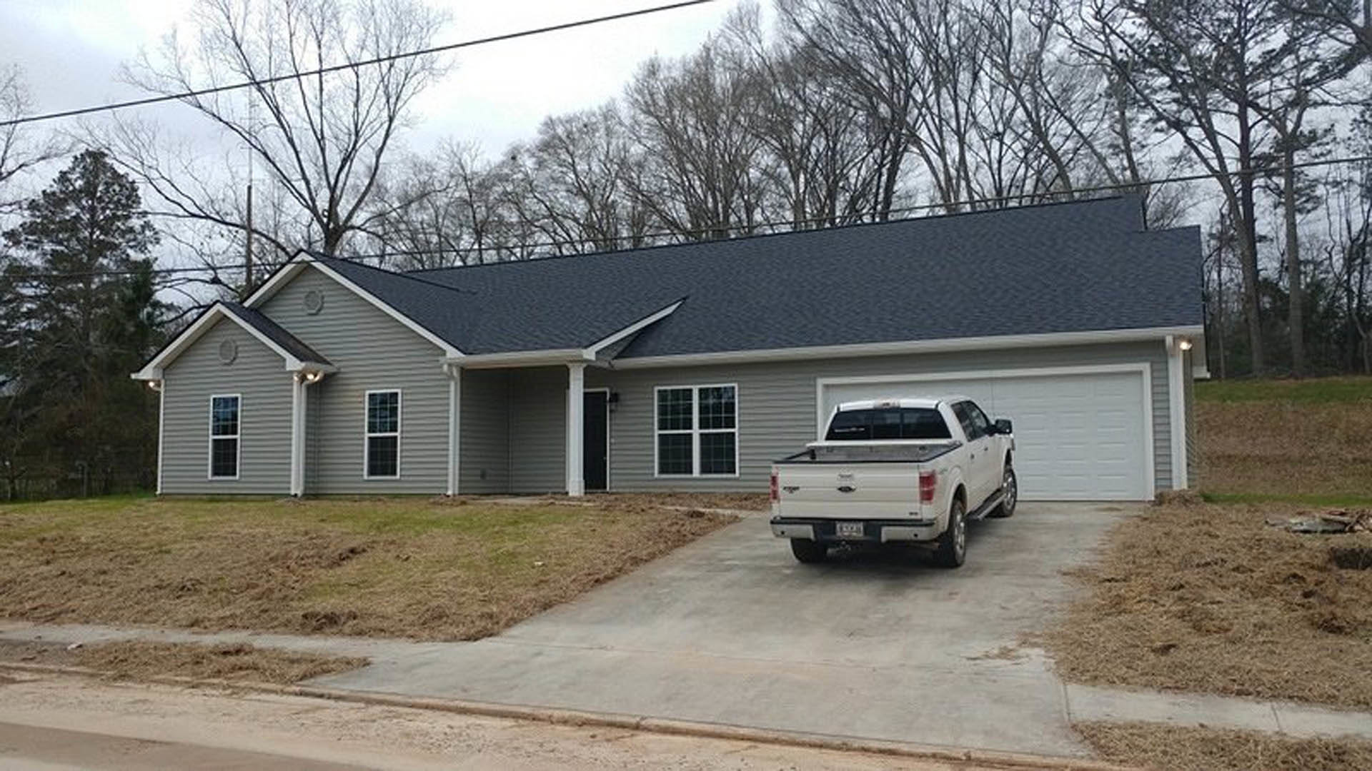 Concrete driveway with a white truck parked in front of a two-story house, multi-pane windows, snow-covered tree, and overcast sky