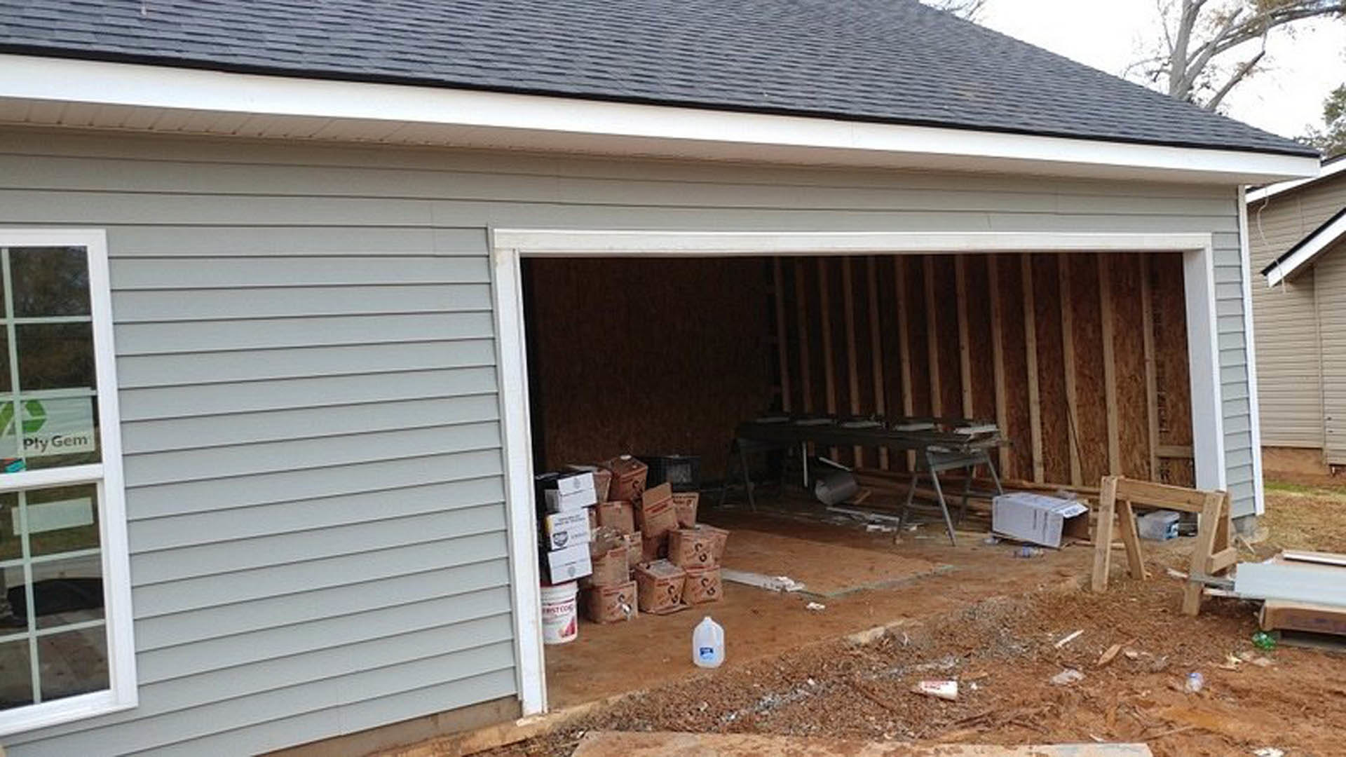 Garage interior with stacked cardboard boxes, white plastic jug with blue label, wood shelving, close-up of window, grey siding, and a backyard shed visible through window.