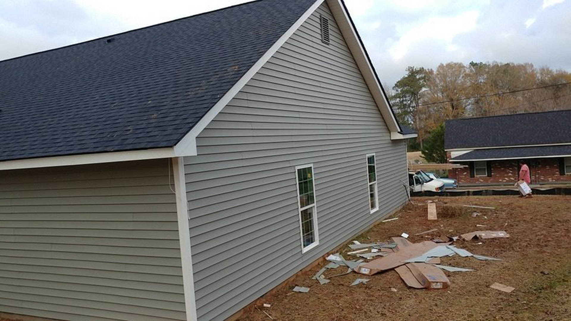 Grey-roofed house with white siding, large pile of cardboard boxes and debris in front yard, single window visible, clear sky overhead